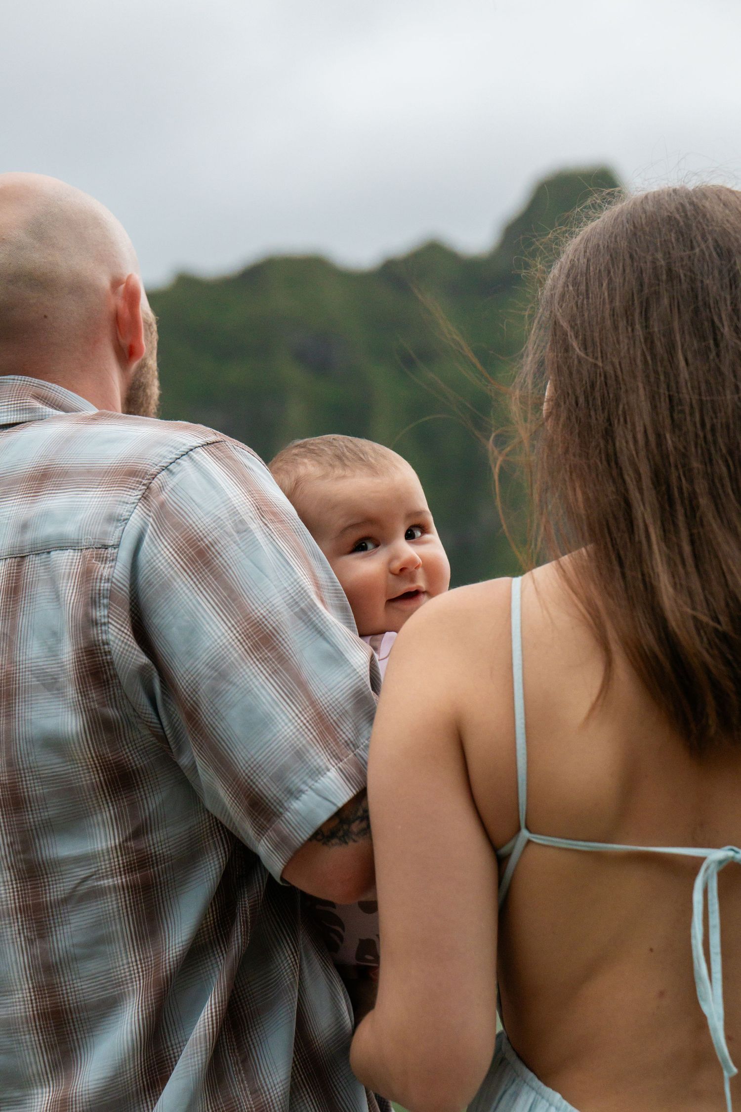 Father holding baby facing lush green Hawaiian mountains while mother stands beside them.