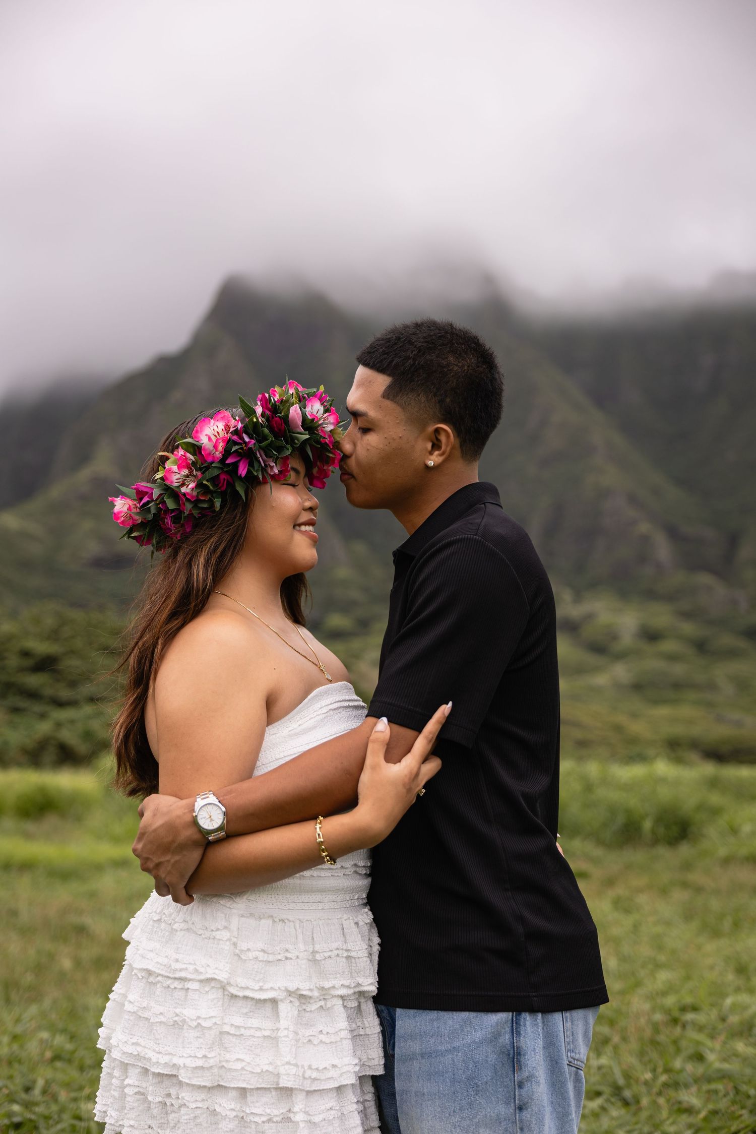 Couple sharing a romantic moment in a lush Hawaiian valley, woman wearing a floral crown and white dress, mountains in background.