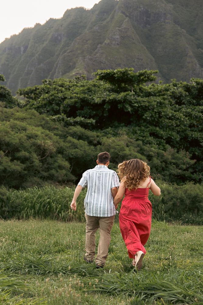 A couple walks hand-in-hand through a green meadow toward lush tropical mountains, woman in a red dress.