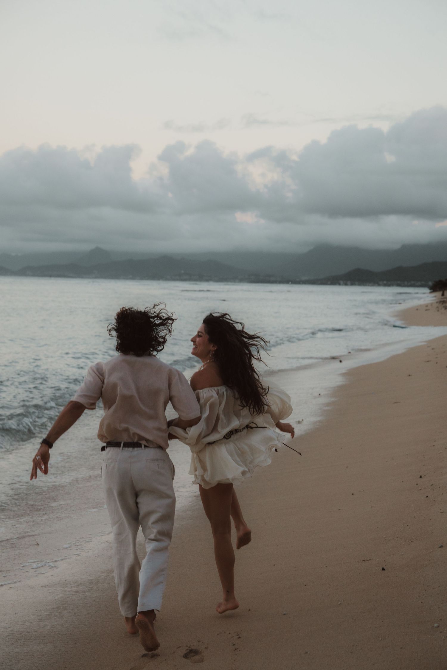 Couple in white outfits runs laughing along sandy beach at dusk.
