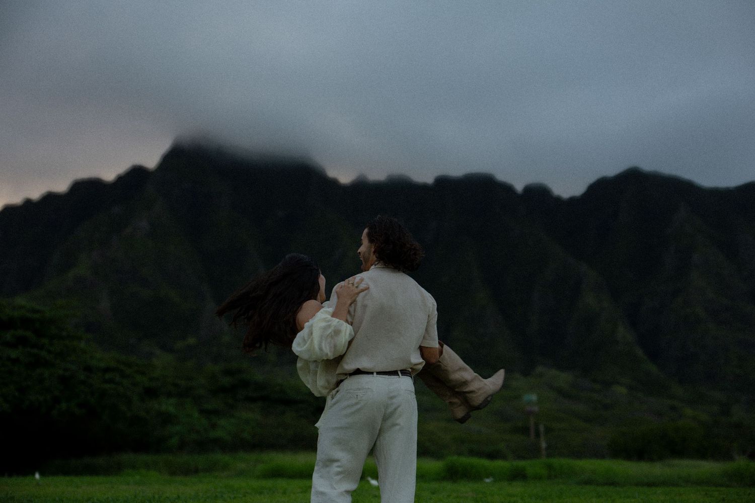 Couple embracing with man lifting woman against moody cloudy Hawaii mountain backdrop at dusk.