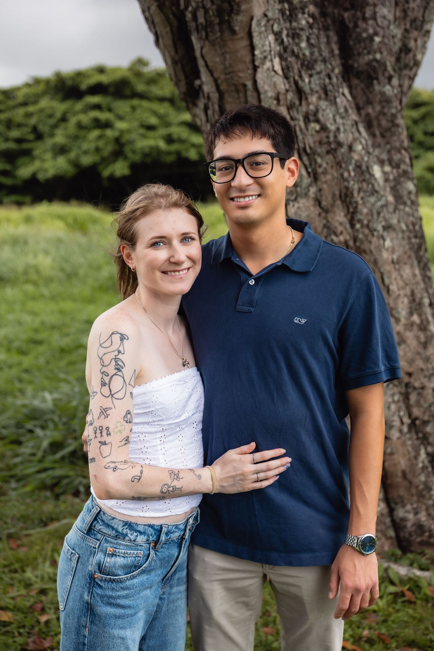 Smiling couple posing outdoors near a large tree, woman with tattoos in white top and jeans, man in navy polo with glasses.