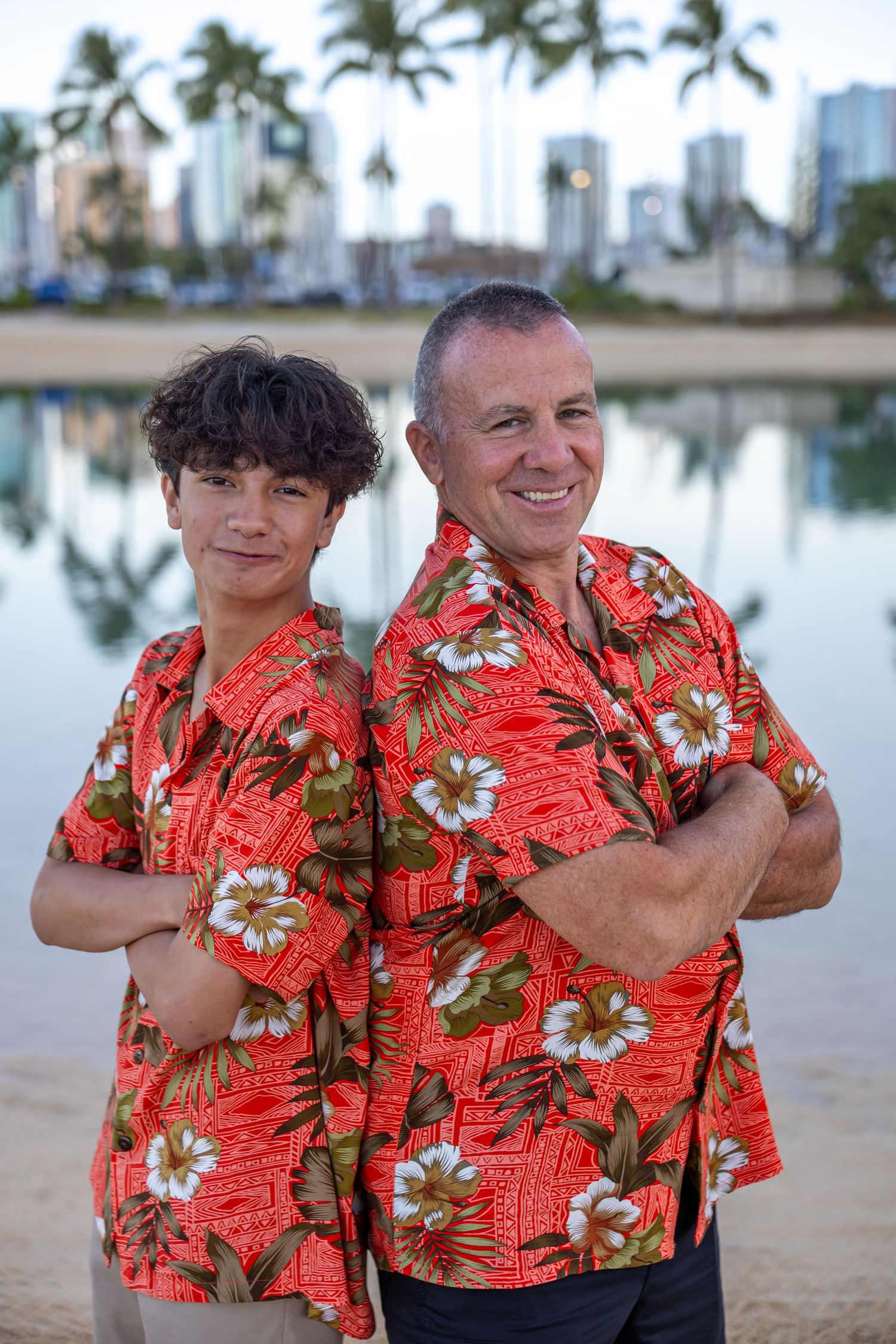 Two men in matching red Hawaiian shirts pose back-to-back by a waterfront with city skyline backdrop.