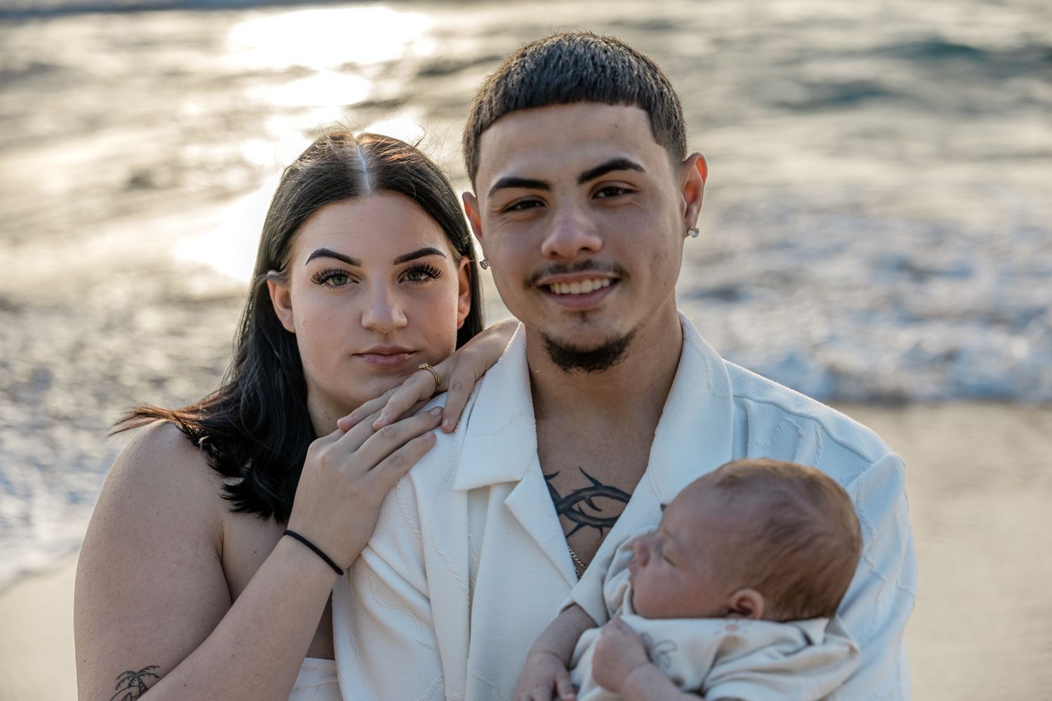 Young couple holding newborn baby on beach at golden sunset hour.
