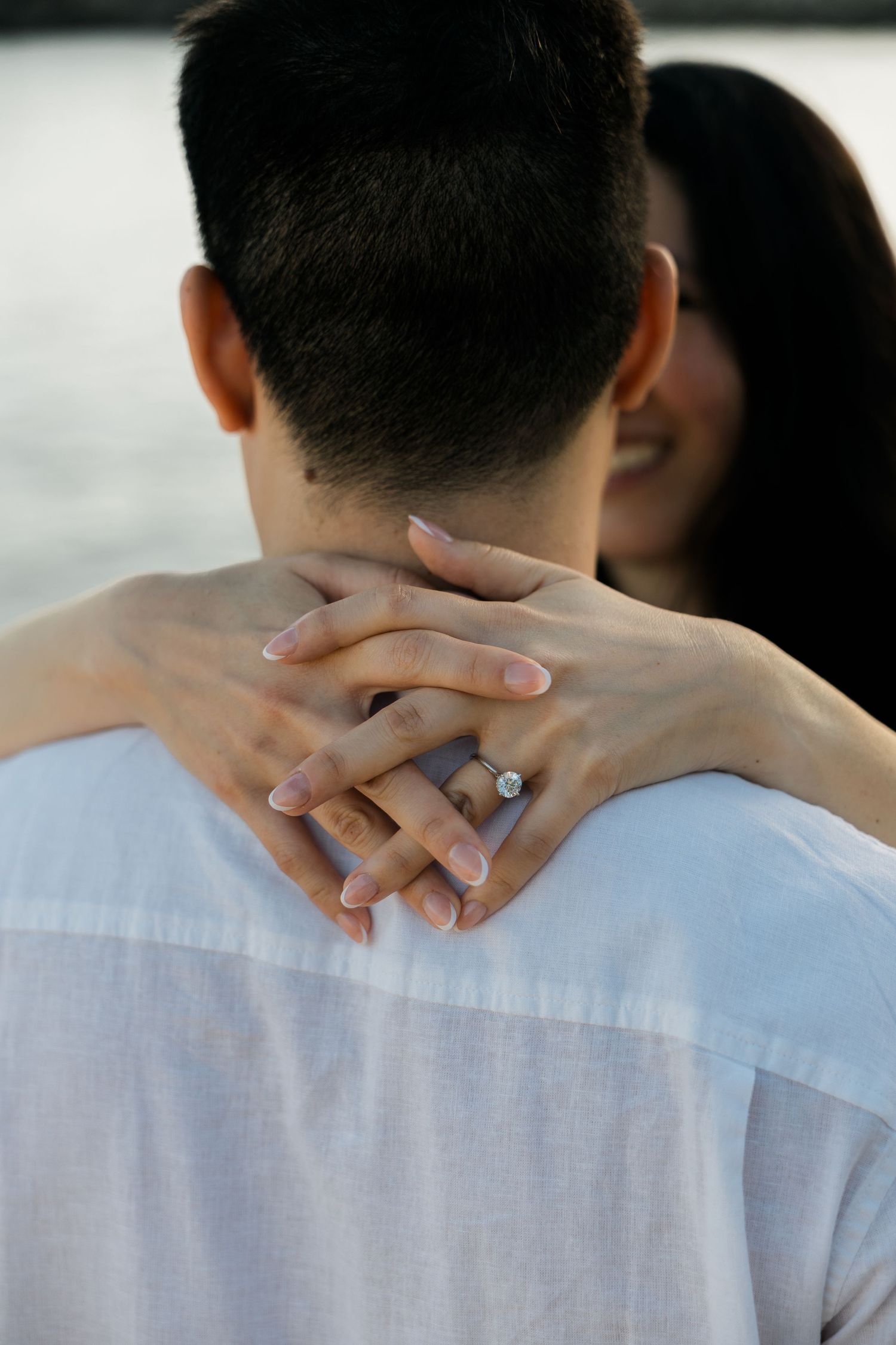 Close-up of womans hands with engagement ring wrapped around mans neck during romantic embrace.