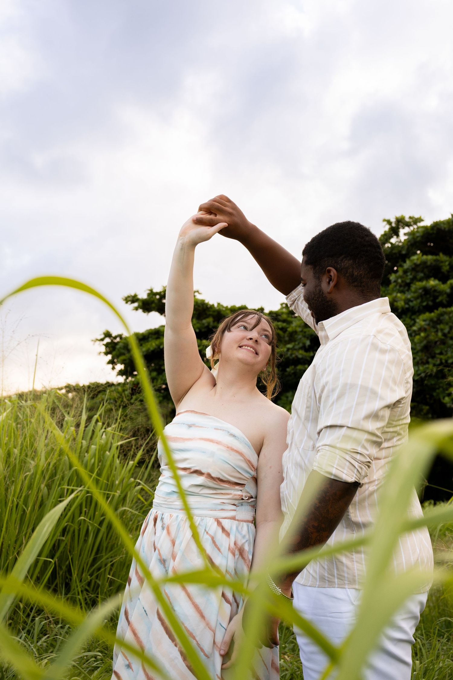 Interracial couple dancing joyfully in a green field at sunset, woman smiling as man twirls her.