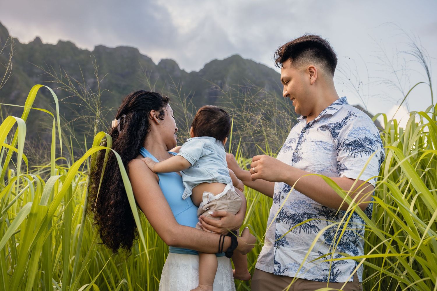 A young Asian family with a baby poses in lush green grass with mountains in the background.