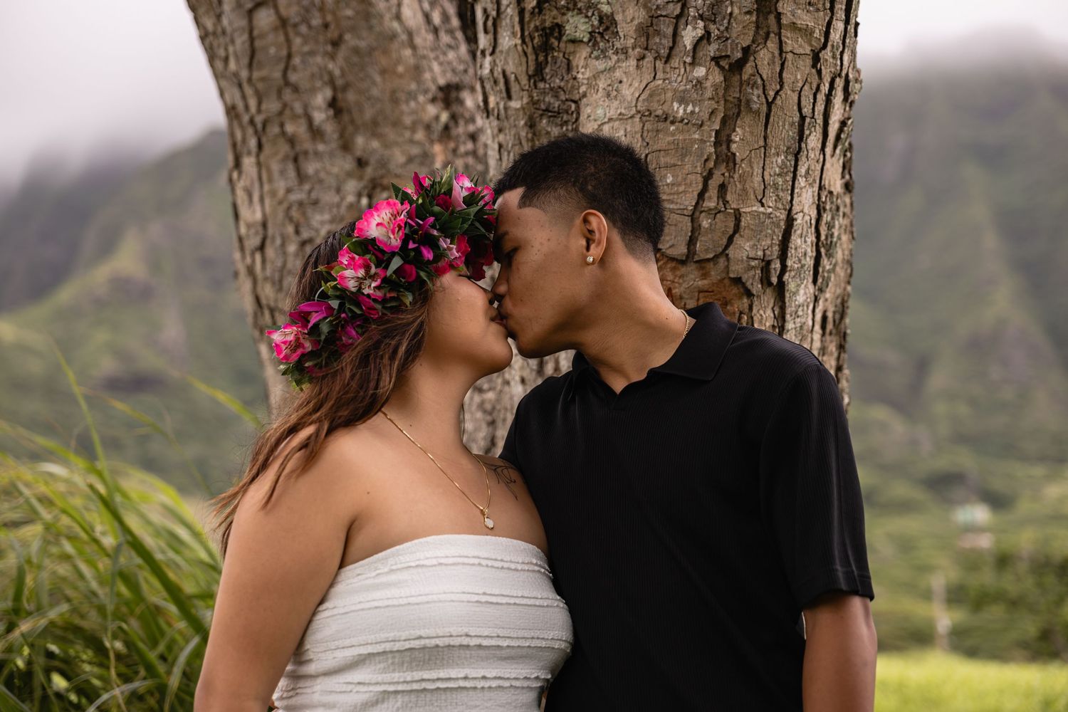 Couple sharing a romantic kiss under a large tree with lush green mountains, woman wearing a pink floral crown.