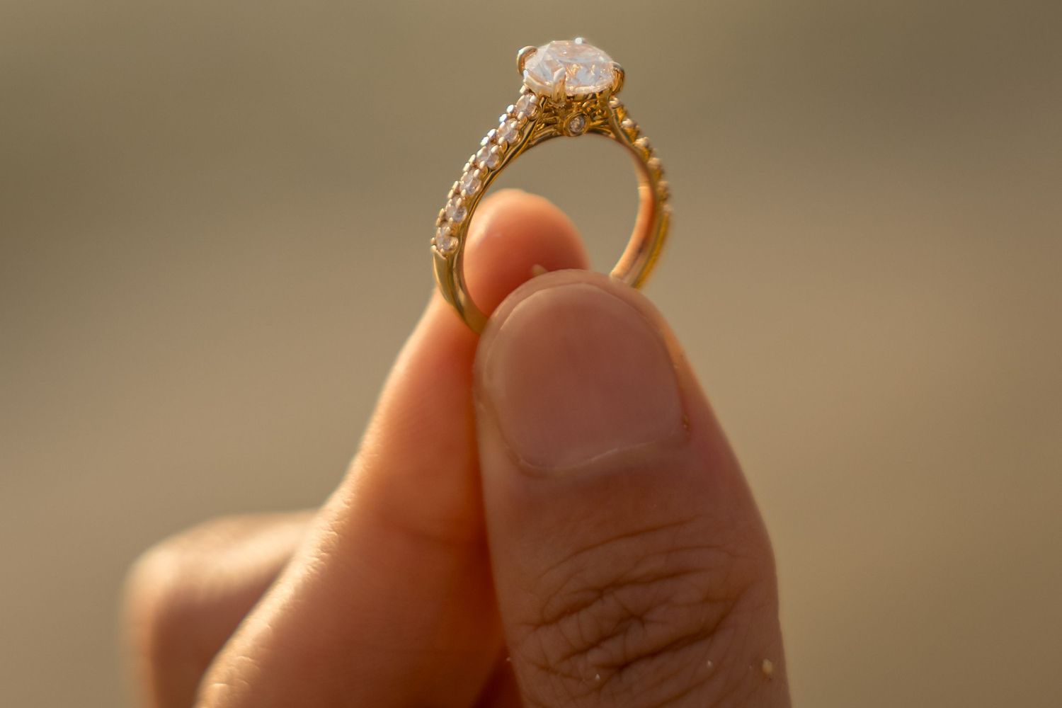 Hand holding a gold diamond engagement ring with intricate band design against a blurred background.
