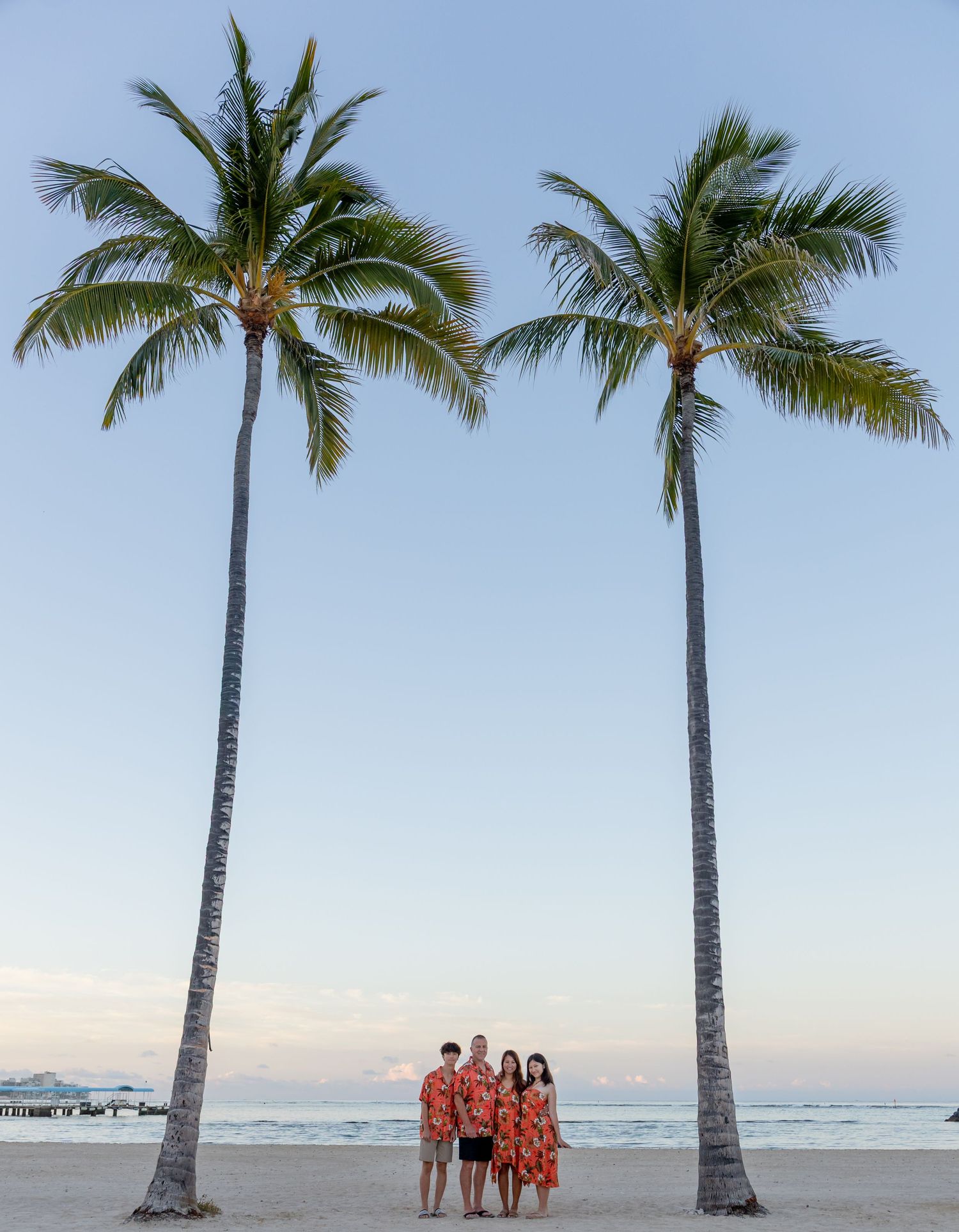 Family of four in matching floral outfits posing between two tall palm trees on a tropical beach at dusk.