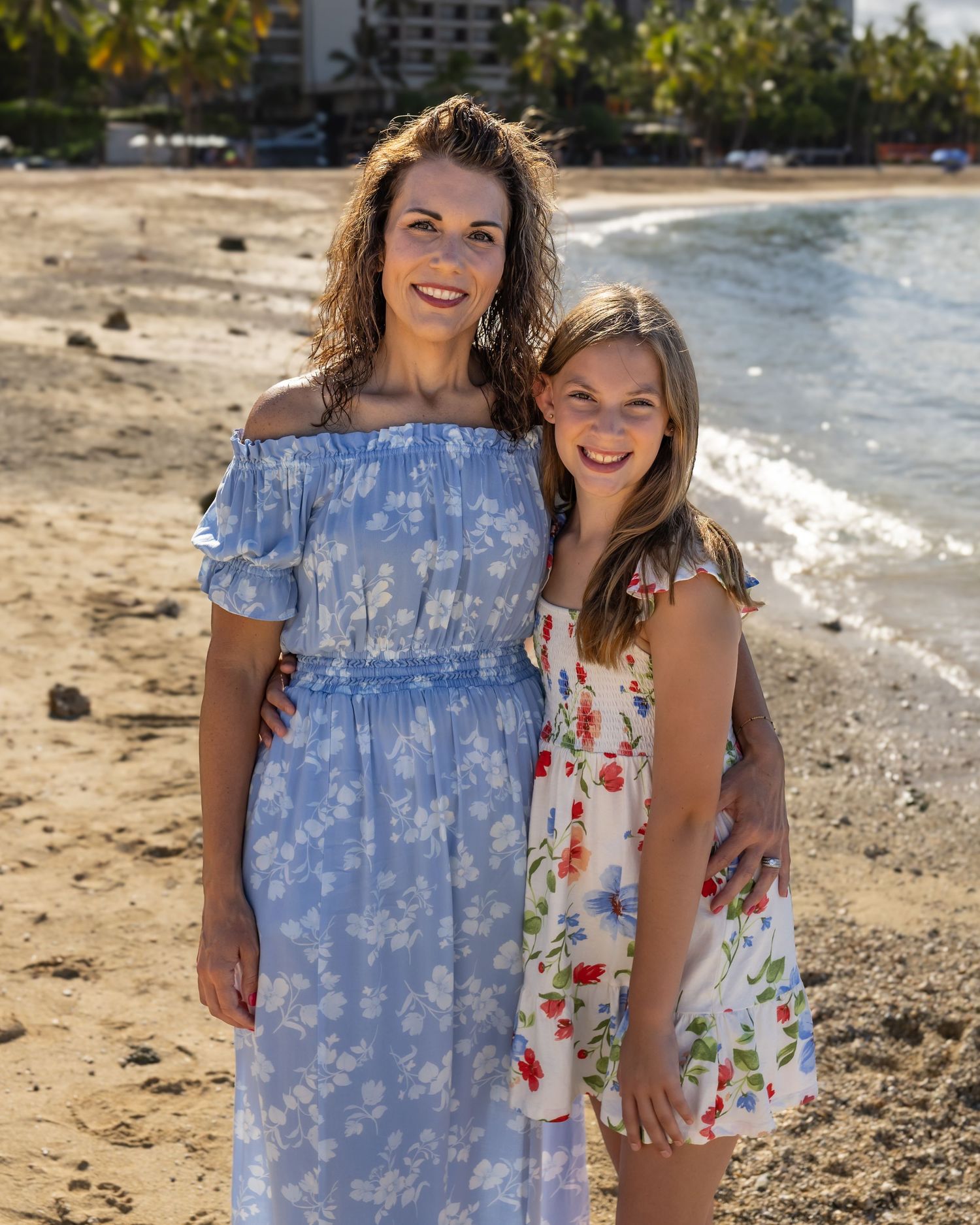 A woman and young girl in floral dresses smile together on a sandy beach with ocean waves behind them.