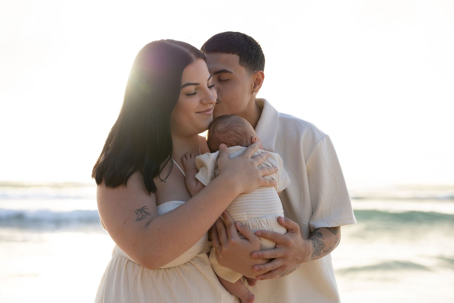 A couple stands on a beach at sunset, tenderly holding and kissing their newborn baby wrapped in a white outfit.