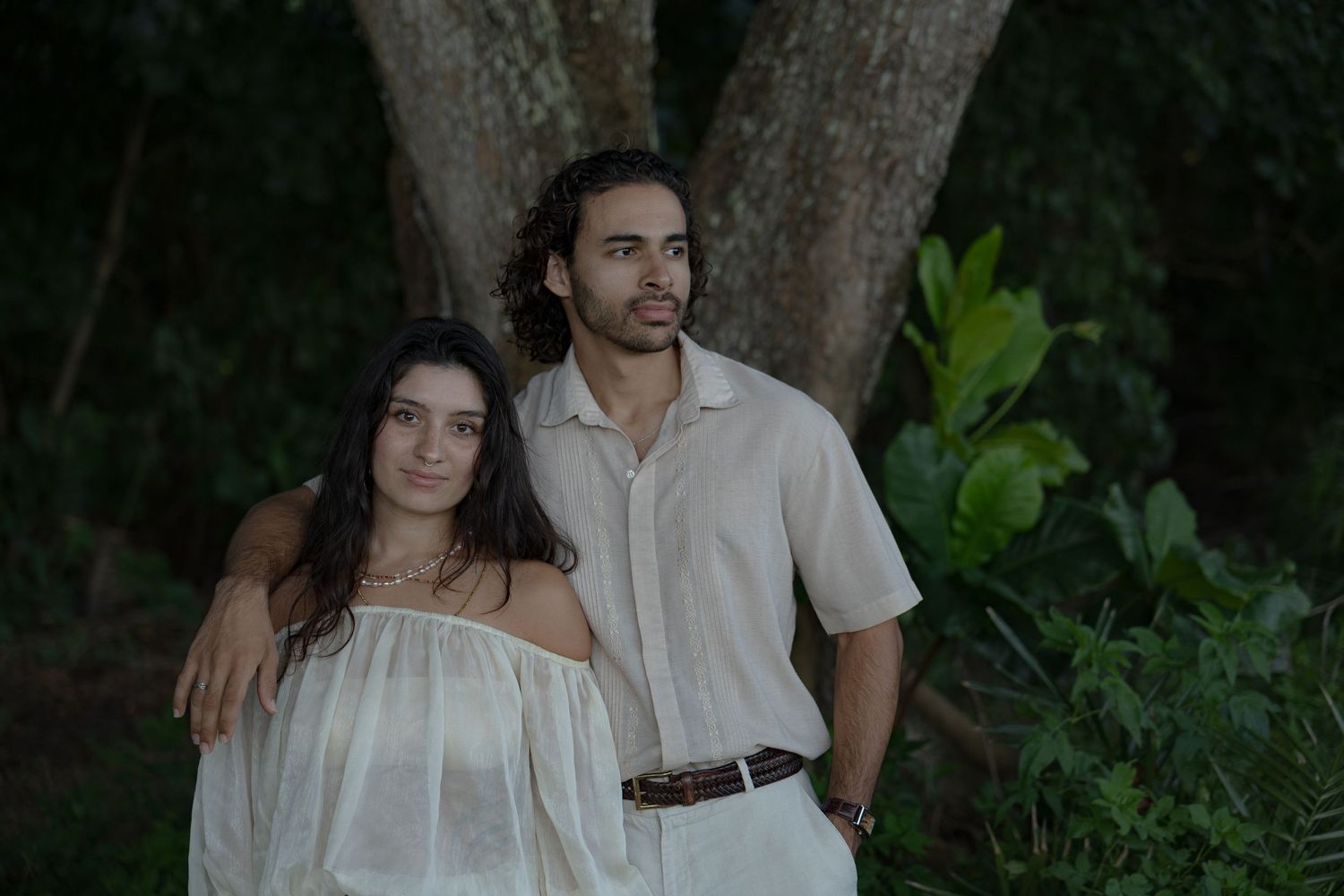 Young couple standing close together in white outfits surrounded by lush tropical forest greenery.