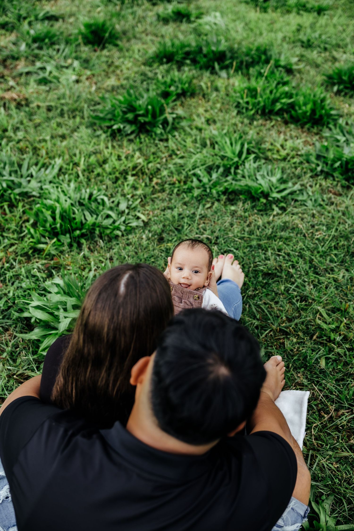 Overhead view of parents sitting on grass watching their smiling baby lying on a blanket.