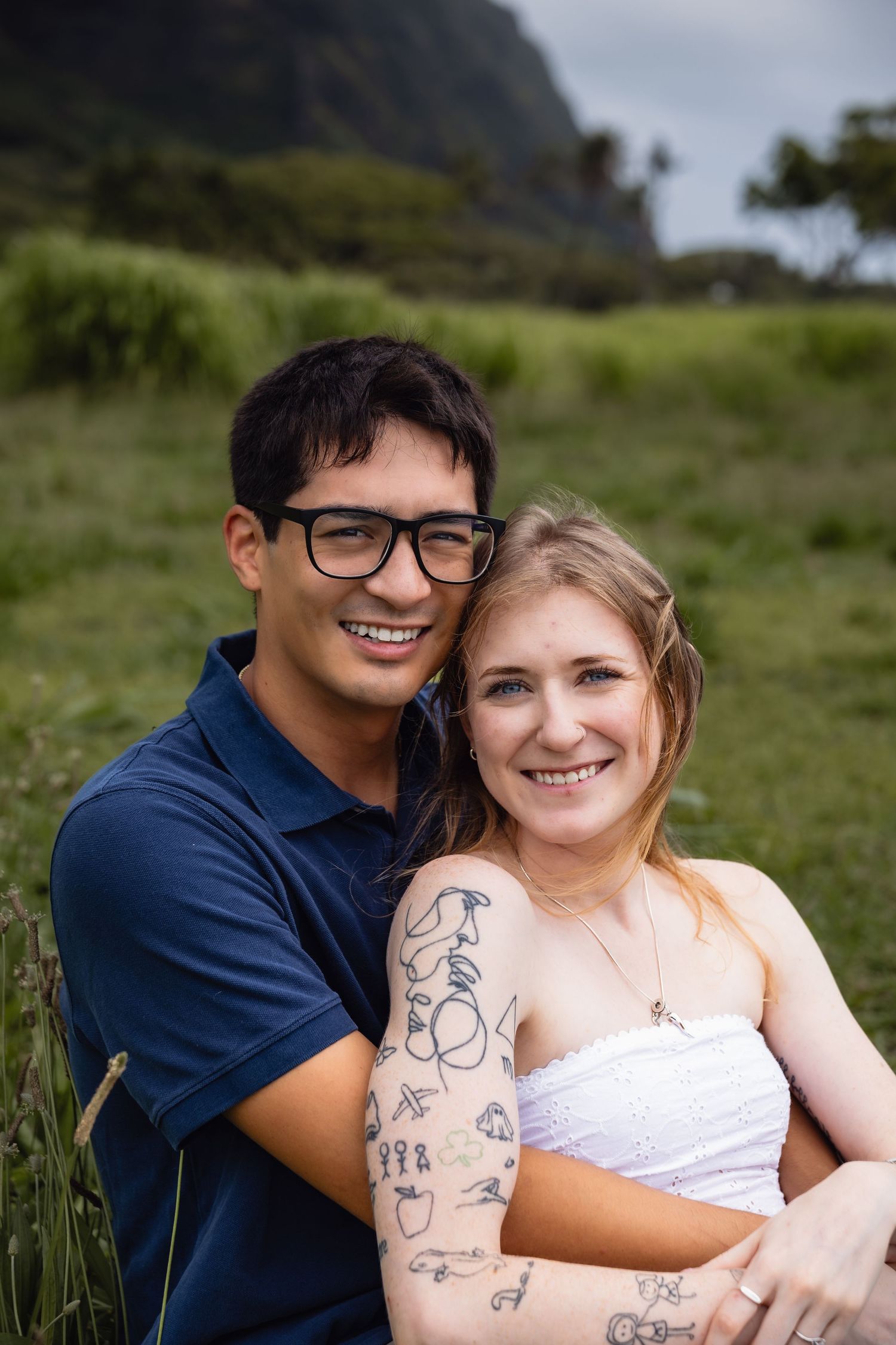 Smiling couple embracing outdoors in a green field, woman with tattoos wearing white top, man in navy polo with glasses.