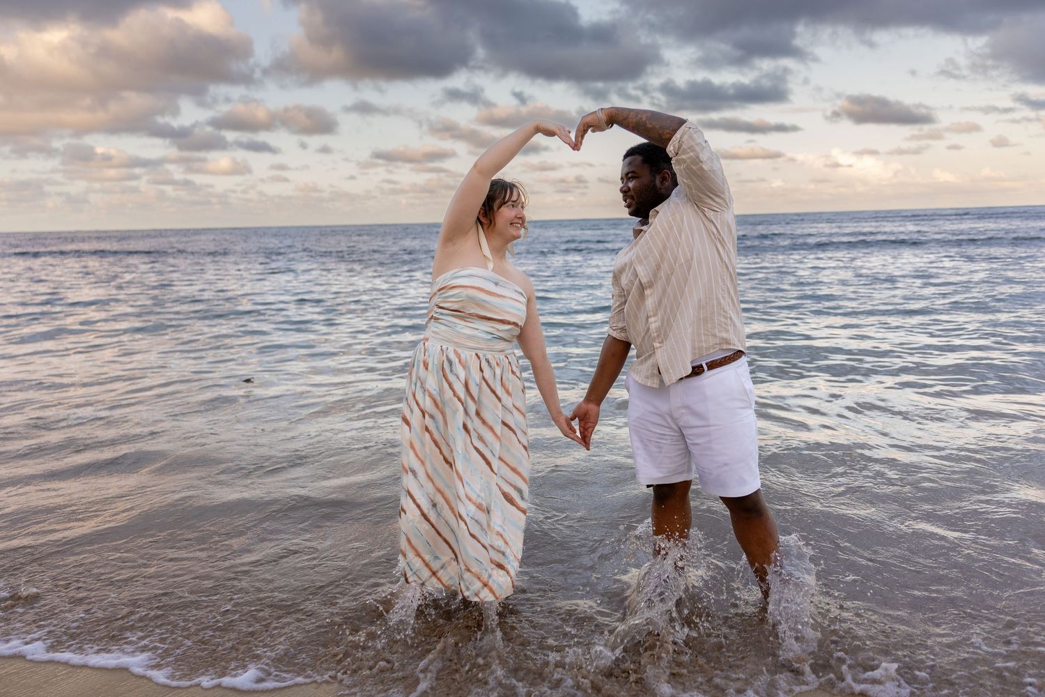 Couple holding hands and making a heart shape with their arms on a beach at sunset with ocean waves.