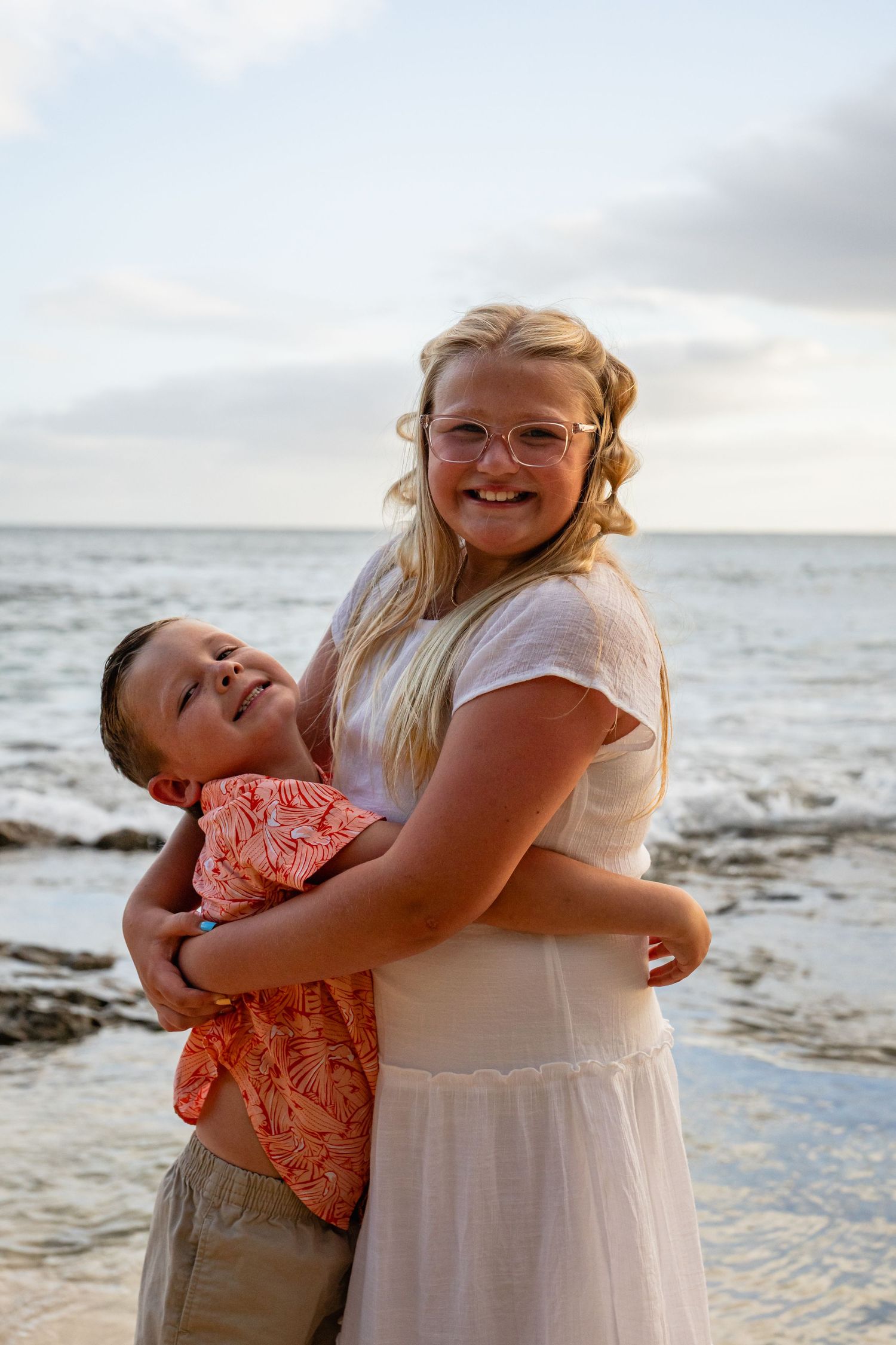 A smiling blonde girl in a white dress hugs a young boy in an orange shirt on a beach at sunset.