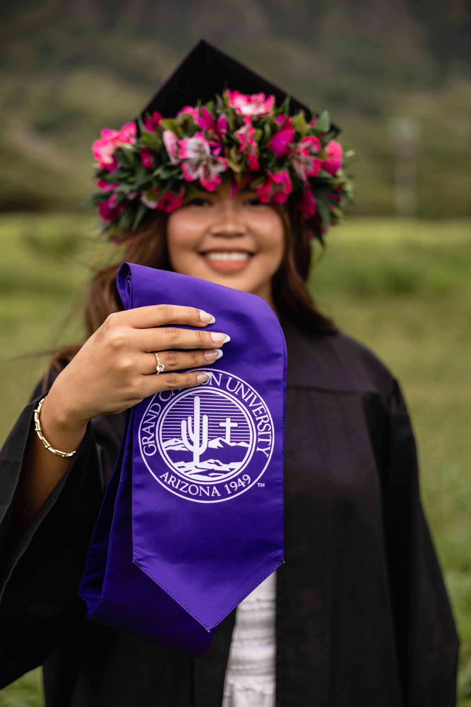 Smiling graduate in black cap with floral lei holds Grand Canyon University purple stole outdoors.