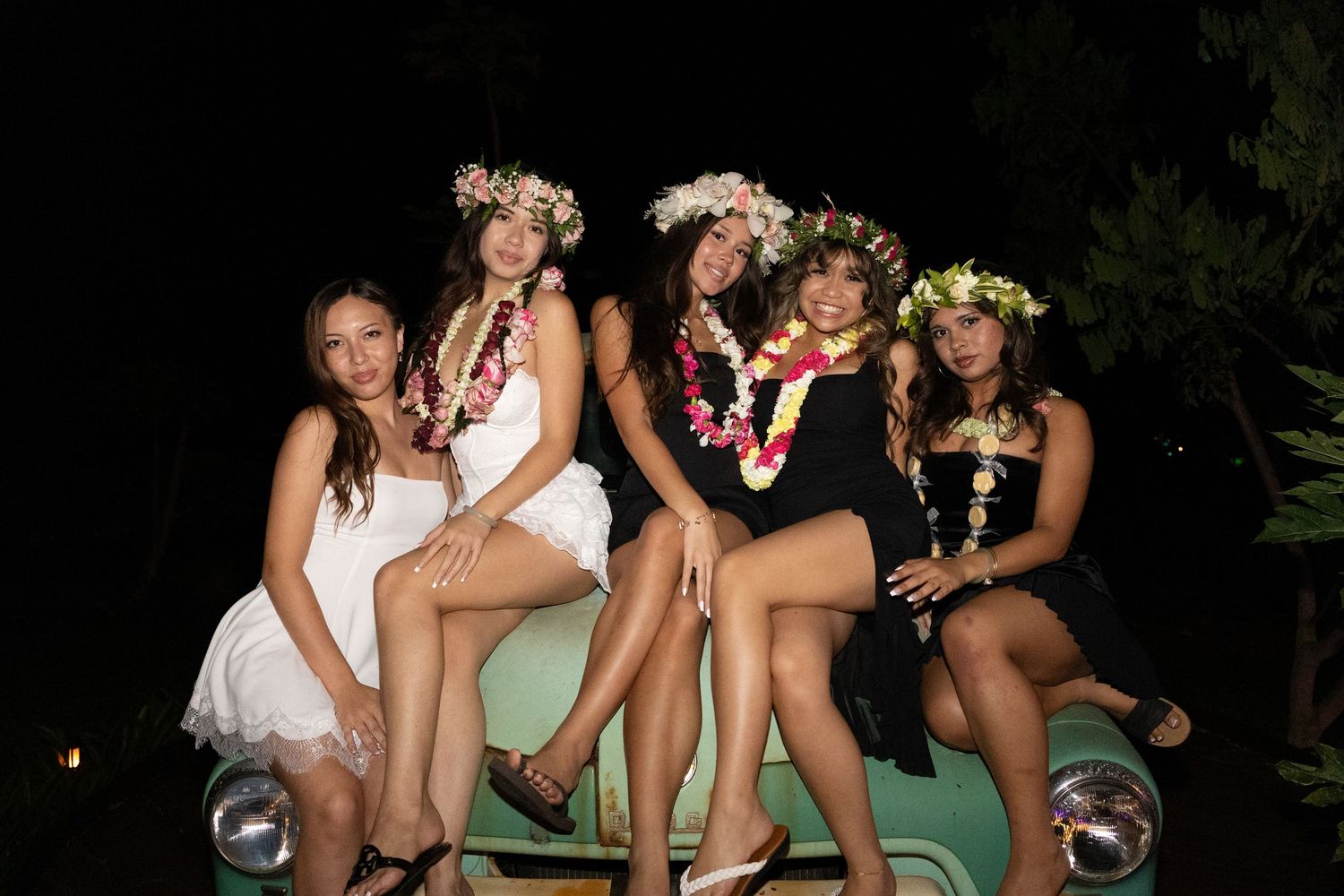 Five young women wearing floral leis and flower crowns pose together on a vintage car at a tropical night event.