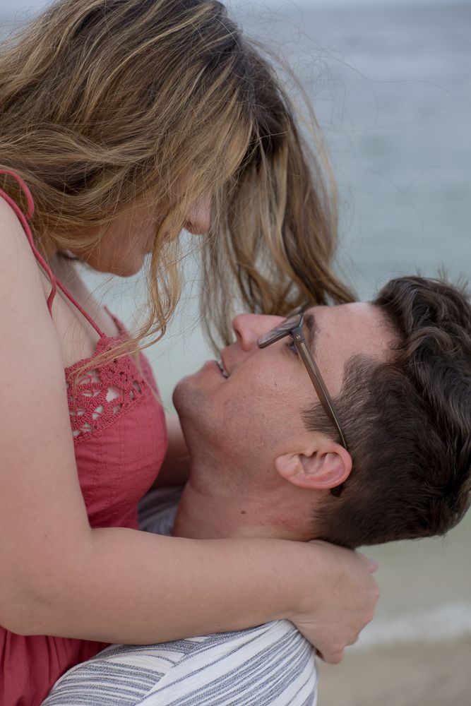 Couple sharing a tender romantic moment on the beach, woman in red dress leaning over man with glasses.