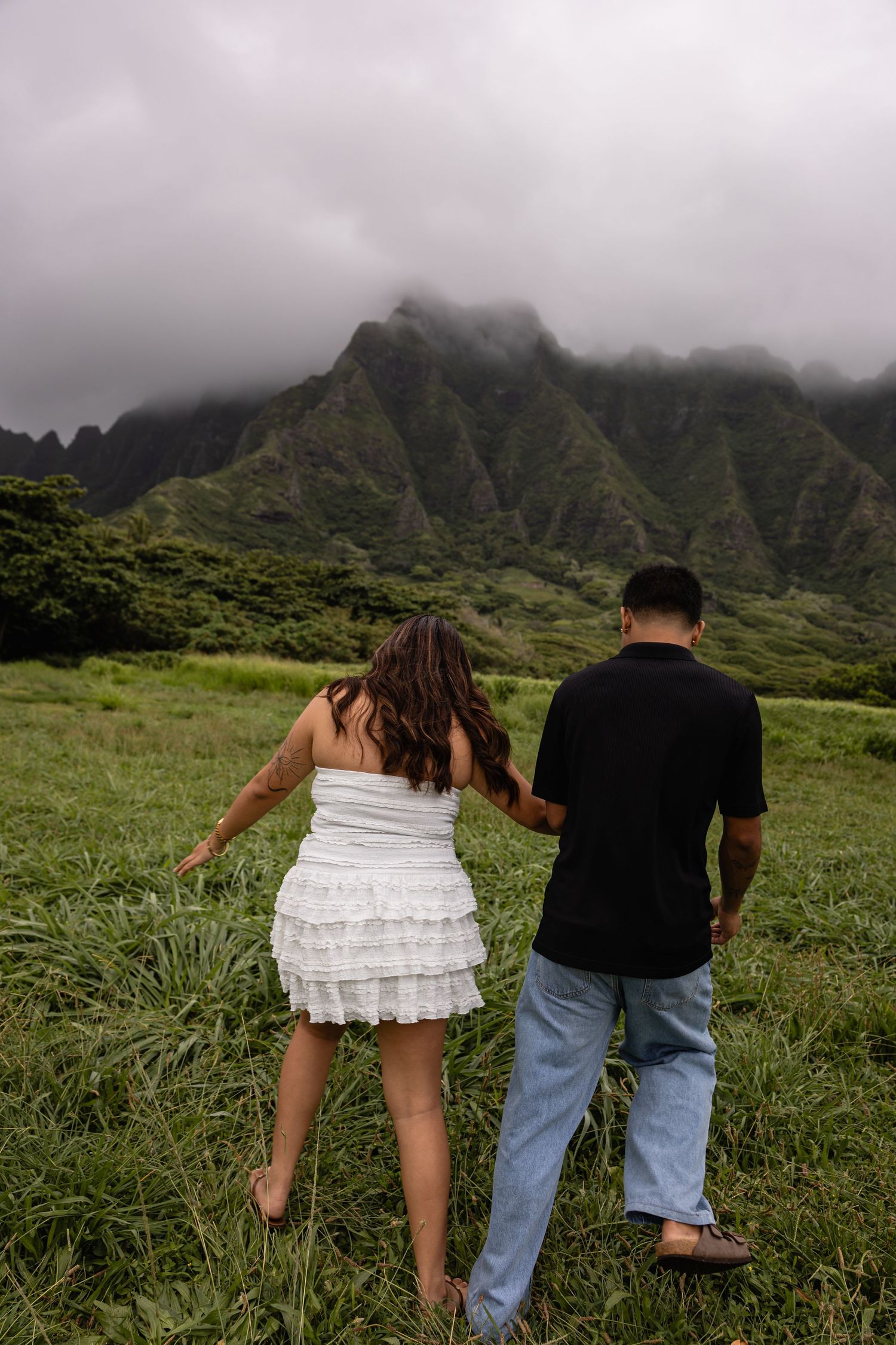 Couple walks hand-in-hand through lush green field toward misty mountain peaks in Hawaii.