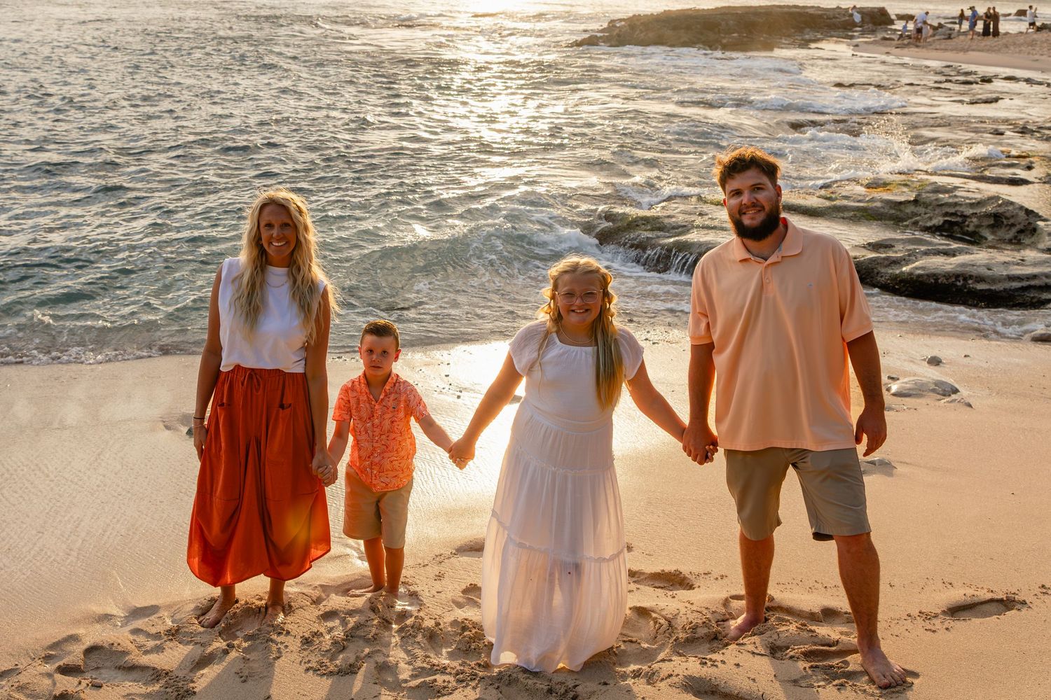Family of four holding hands on sandy beach at golden sunset with ocean waves and rocks in background.