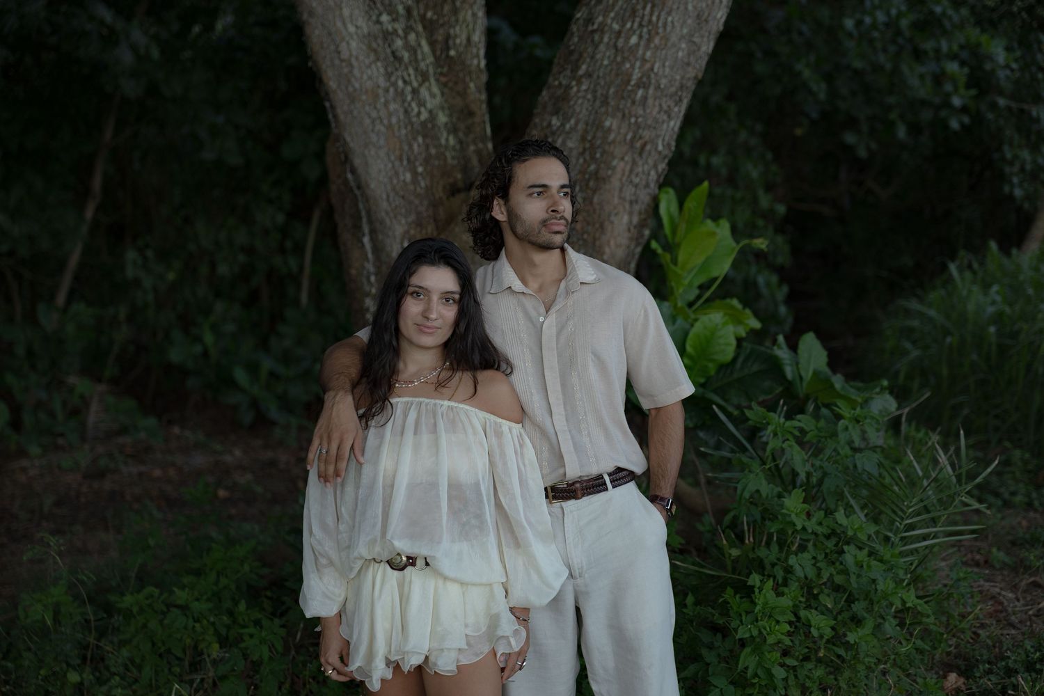 Couple in white outfits posing together in front of large tropical trees in Hawaii.
