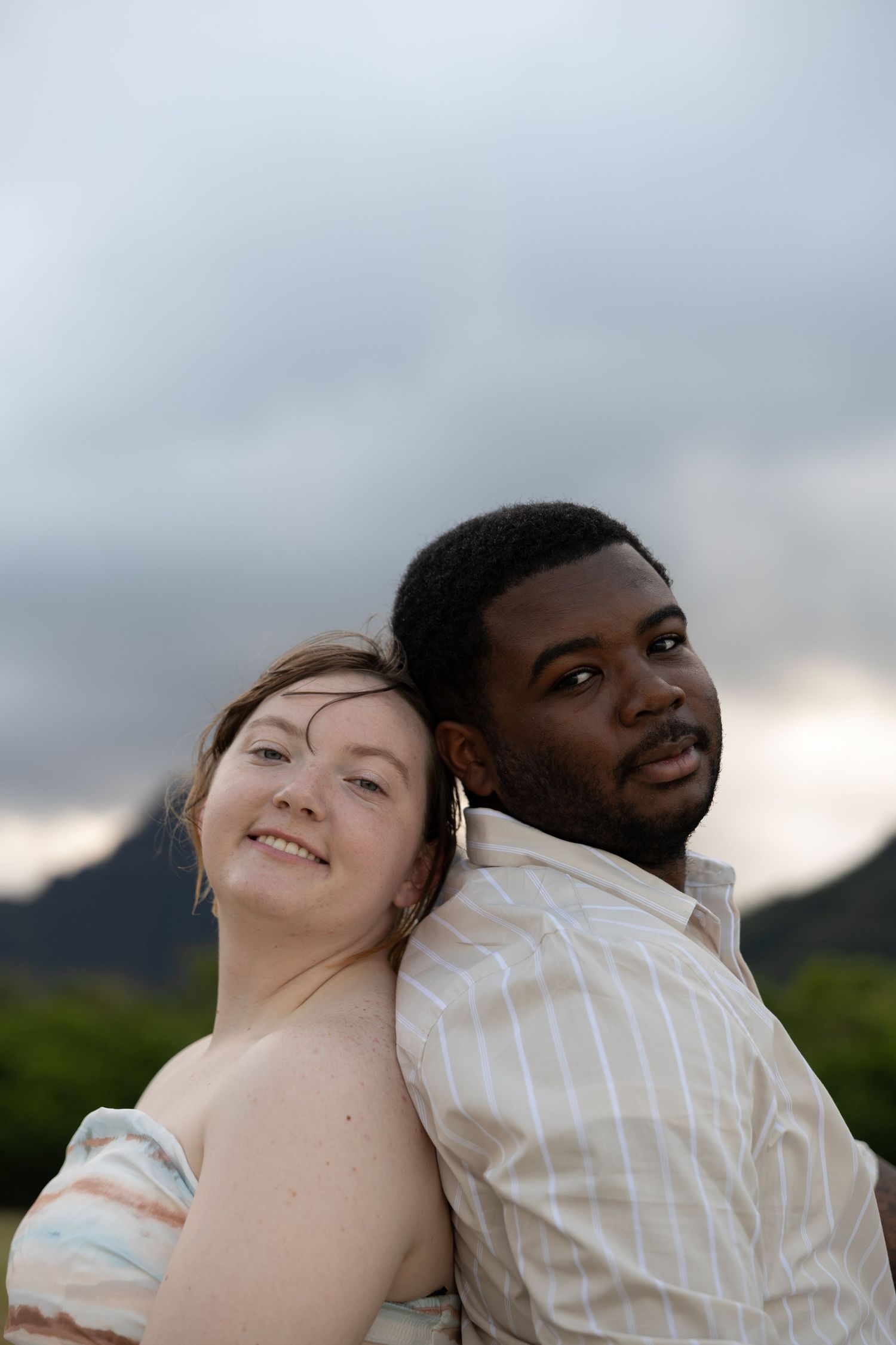 Smiling interracial couple posing back-to-back outdoors with cloudy mountain scenery behind them.