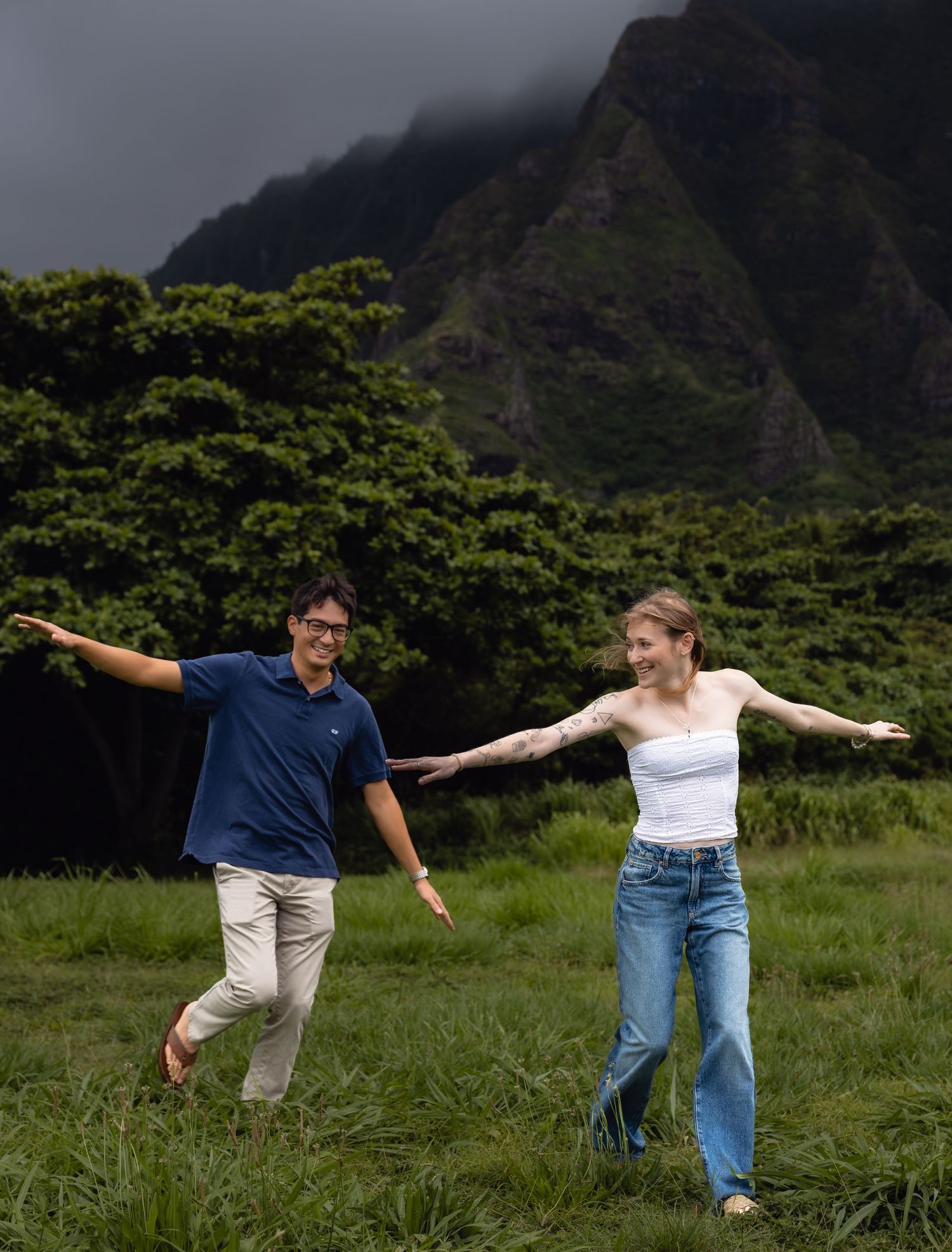 Couple joyfully balancing with arms outstretched in a lush green field with dramatic misty mountains in Hawaii.