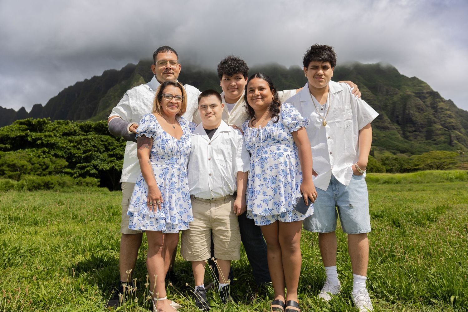 Family of six smiling together in matching blue floral and white outfits with dramatic Hawaii mountains backdrop.