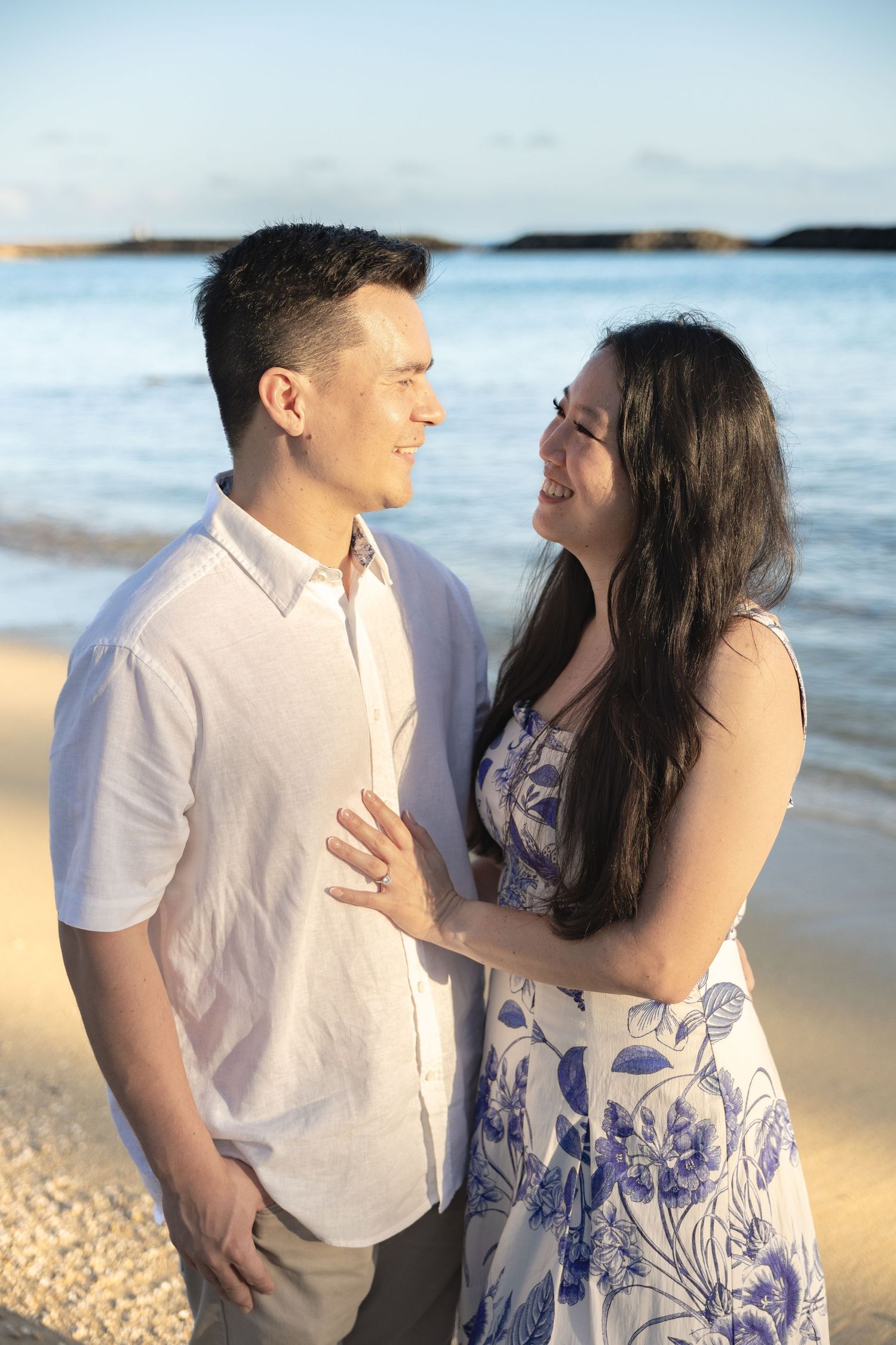 Smiling couple gazing at each other on sandy beach with blue ocean waves behind them.