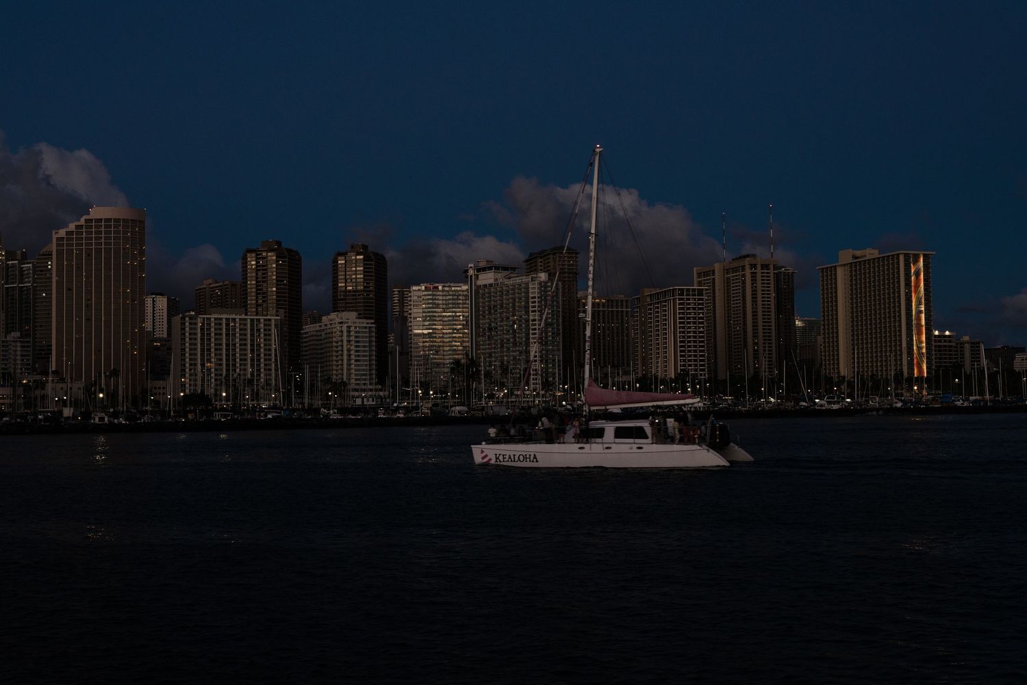 Waikiki city skyline at night with a sailboat on dark harbor waters in foreground.