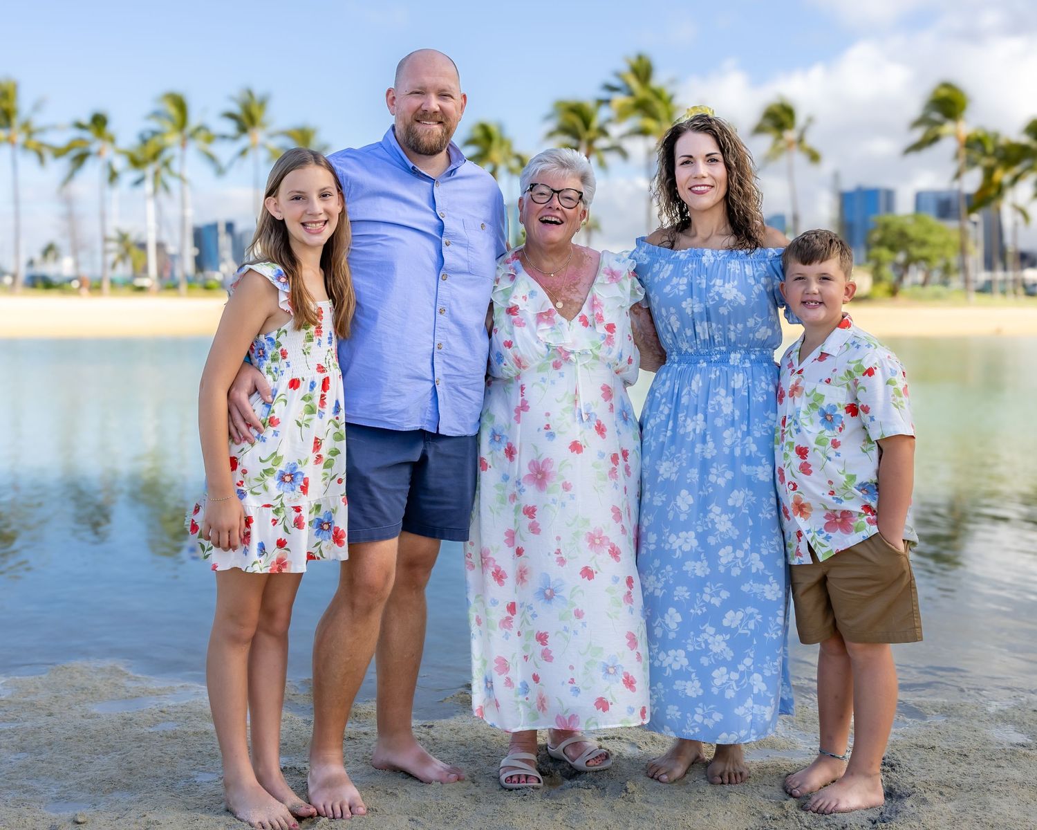 Family of five smiling together on a tropical beach with palm trees and city skyline in the background.