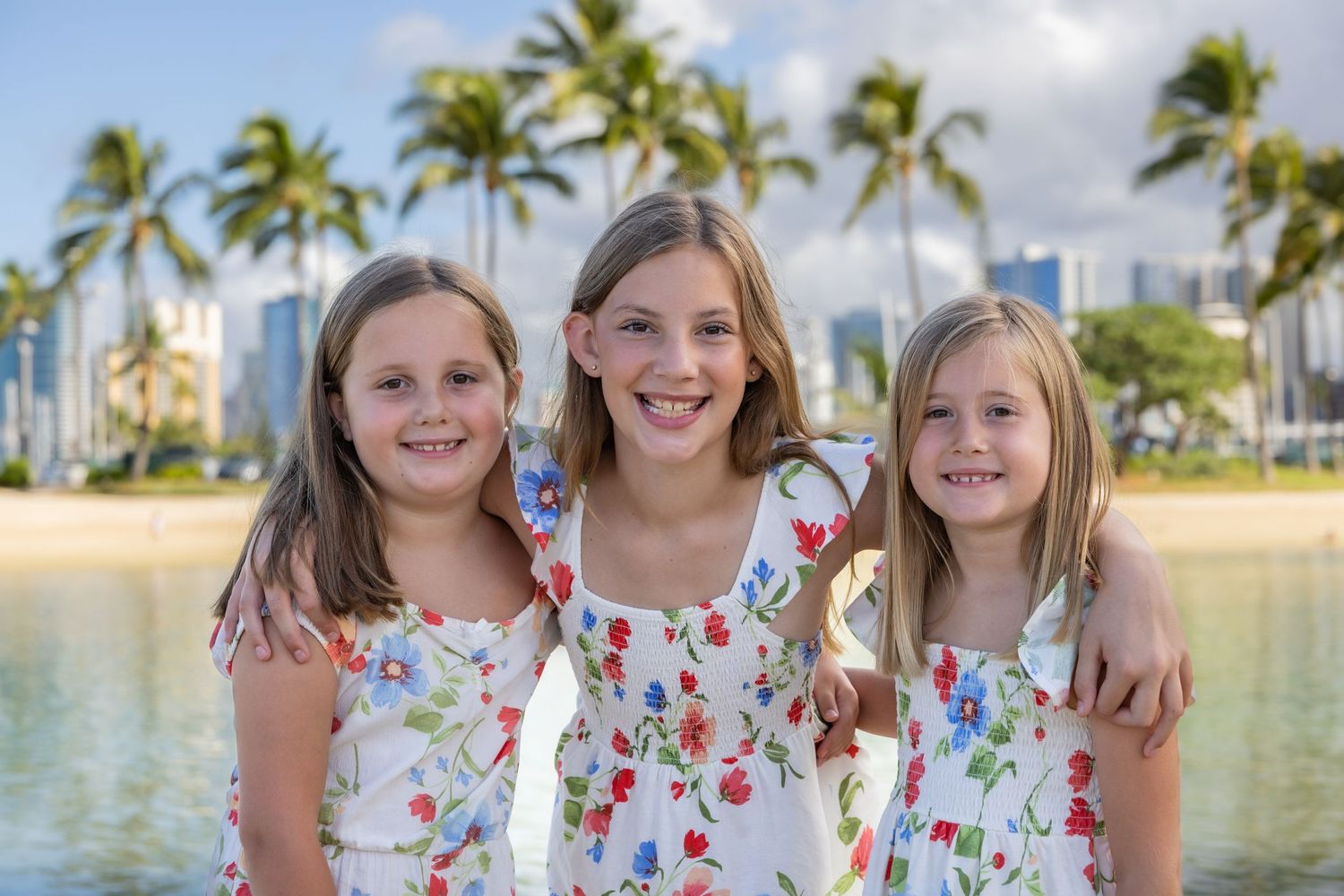 Three young girls in matching floral dresses smile together on a Hawaii beach with palm trees and city skyline.