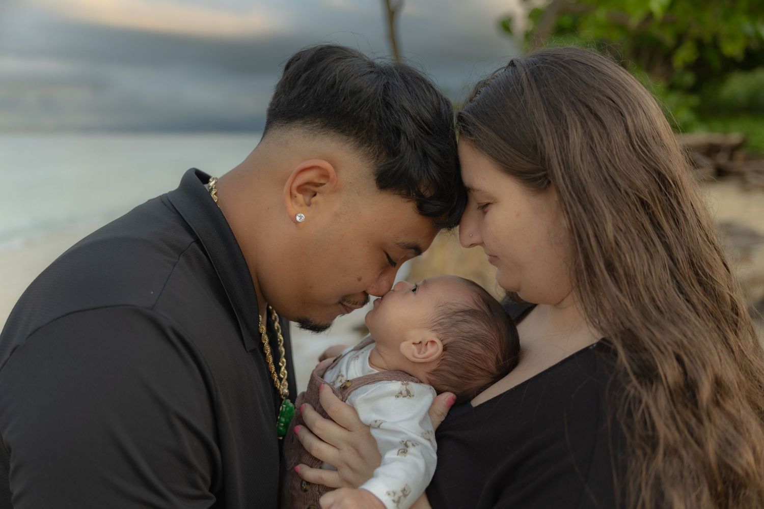Loving parents touching foreheads with newborn baby on Hawaii beach at dusk.