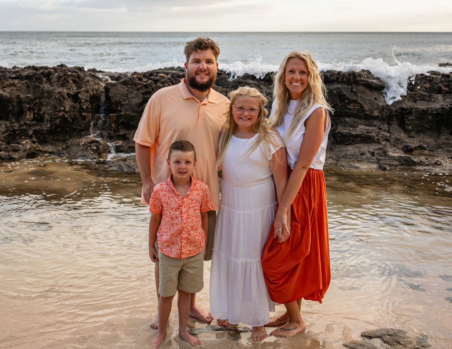 Family of four smiling together on a rocky beach at sunset, wearing coordinated outfits in warm tones.