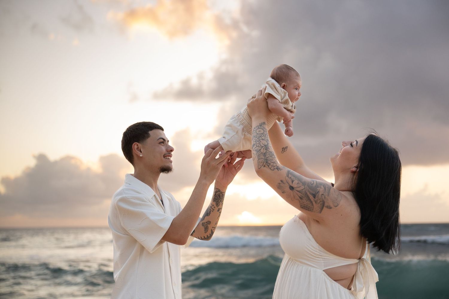 Tattooed couple lifting newborn baby at sunset on ocean beach in white outfits.