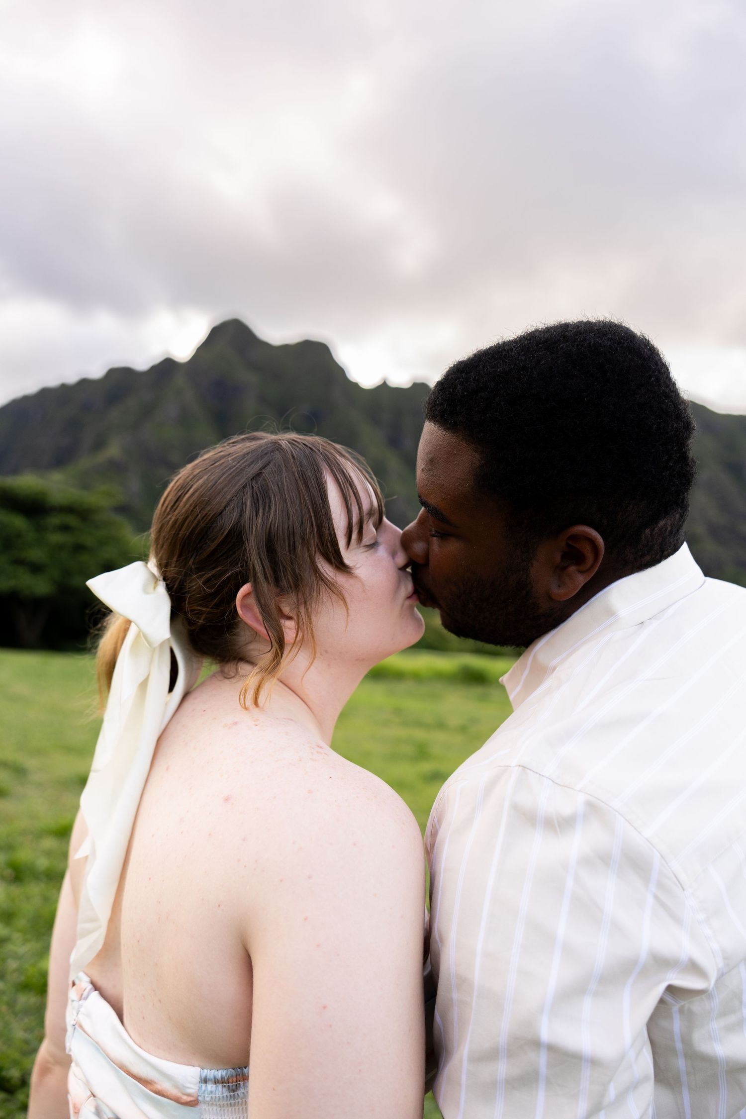Interracial couple sharing a kiss in a lush green field with dramatic mountain backdrop in Hawaii.