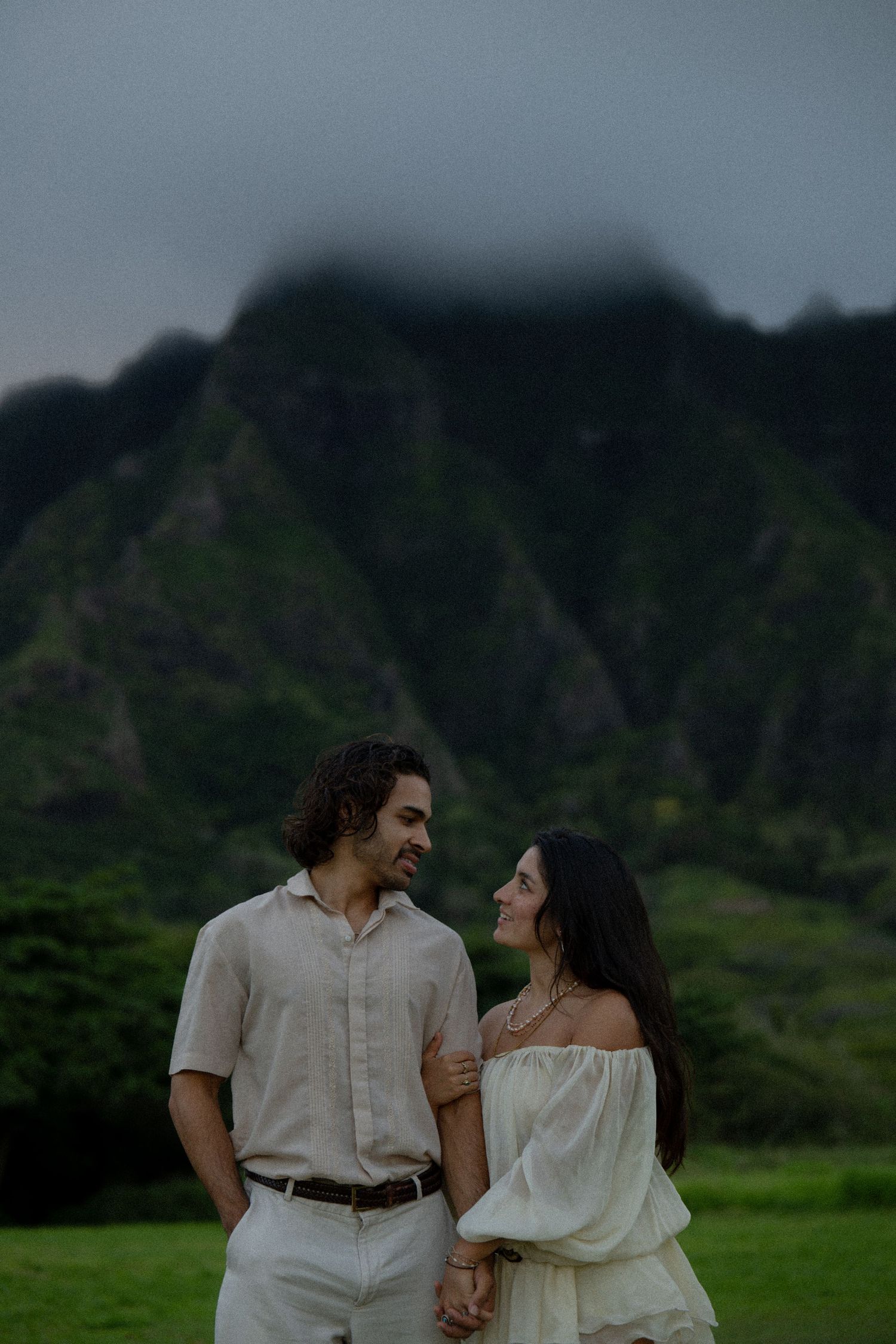 Couple holding hands smiling at each other with misty green mountains behind them.