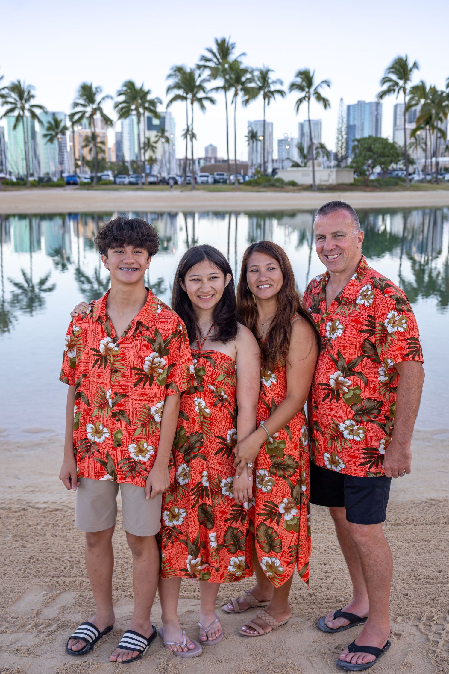 Family of four in matching red Hawaiian outfits posing on a sandy beach with palm trees and city skyline reflected in calm water.