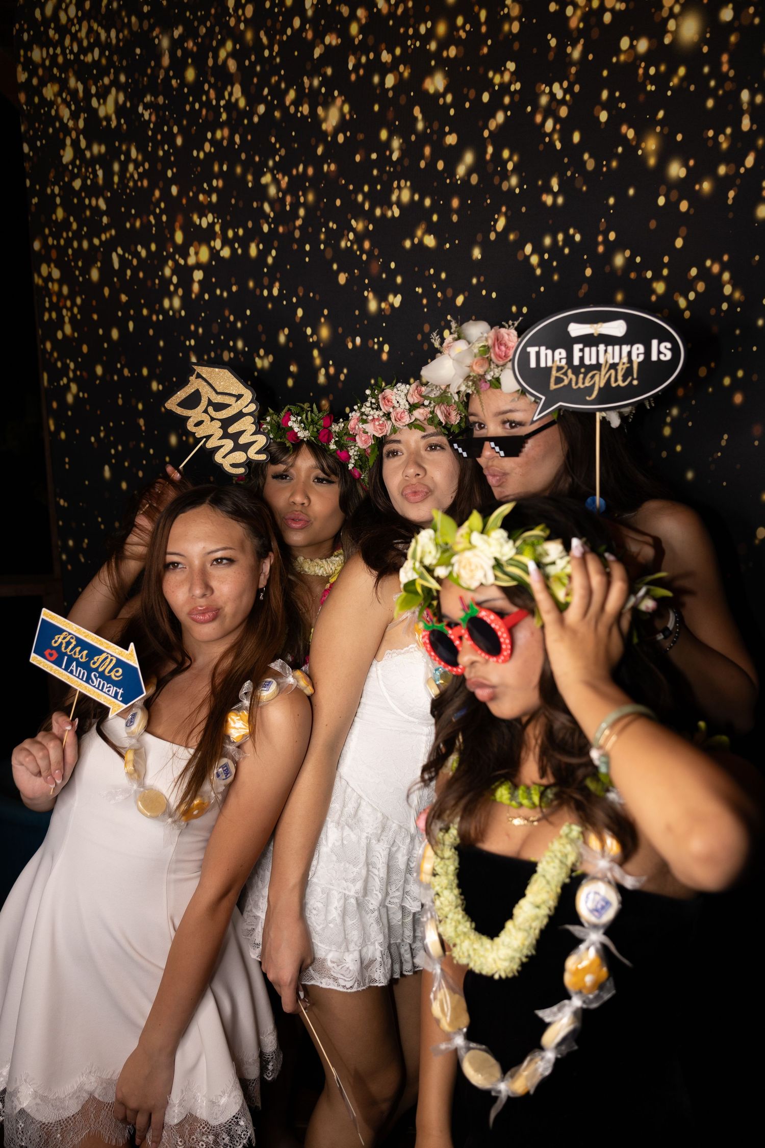 Group of young women posing in a photo booth with floral leis, props, and a sparkly black backdrop.