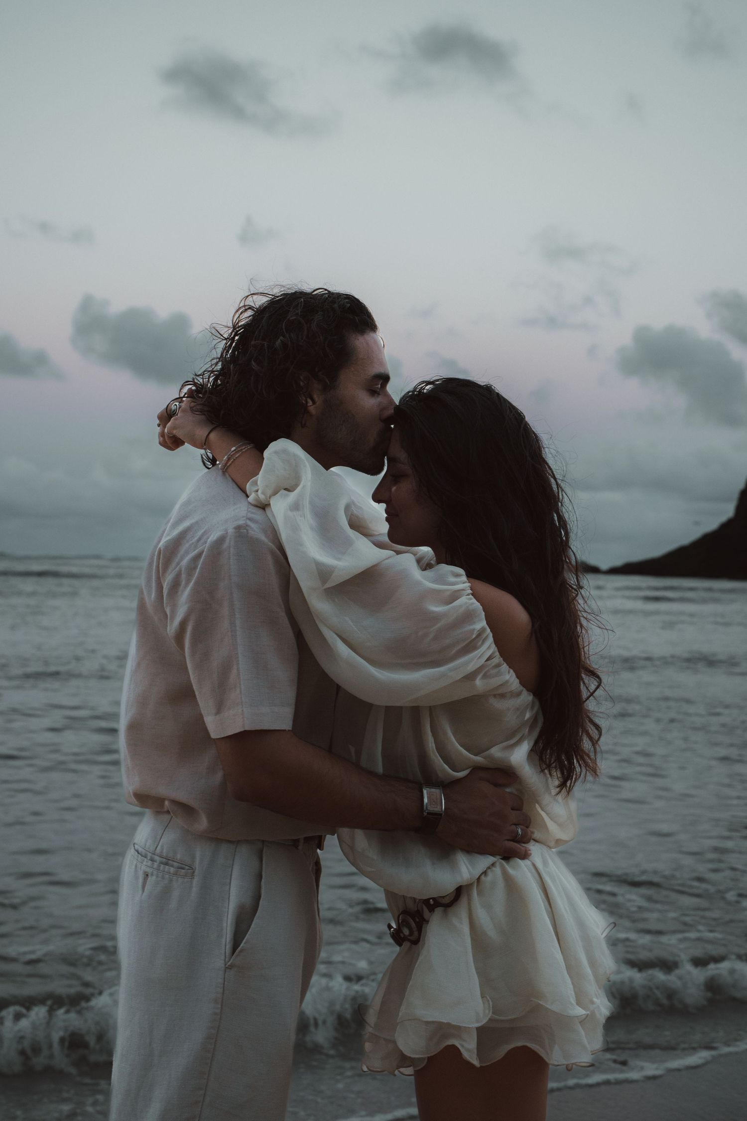 Romantic couple sharing a kiss on a Hawaii beach at sunset, both dressed in elegant white outfits.
