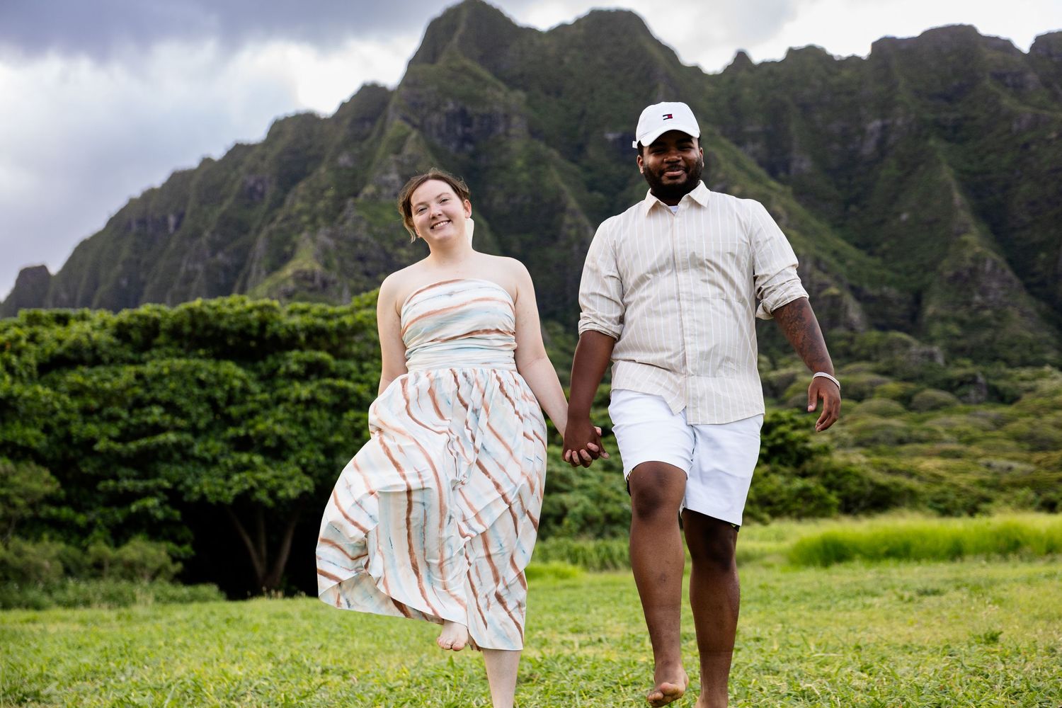 Couple holding hands walking on green grass with dramatic Ko'olau Mountains backdrop in Hawaii.