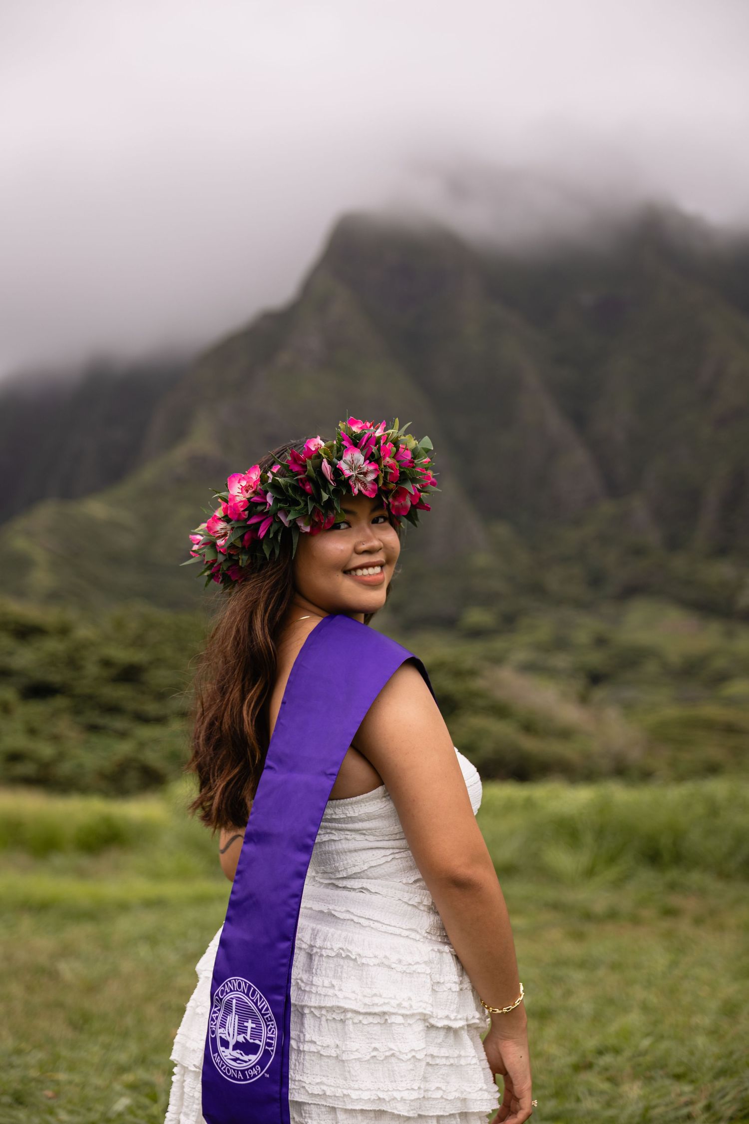 Smiling graduate in white dress with purple sash and floral crown poses before lush green Hawaiian mountains.