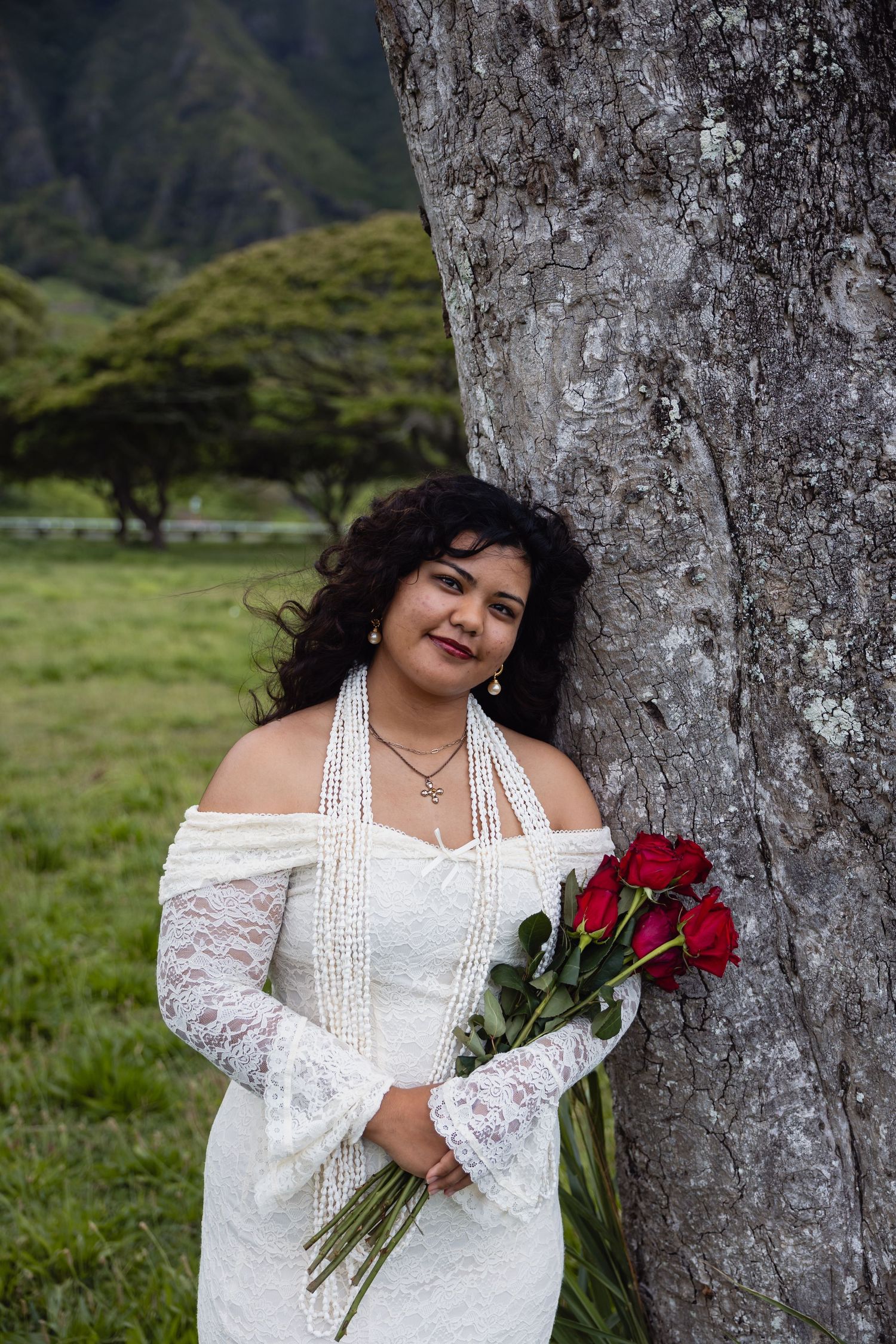Bride in white lace off-shoulder gown holding red roses, leaning against tree with green mountains in Hawaii.