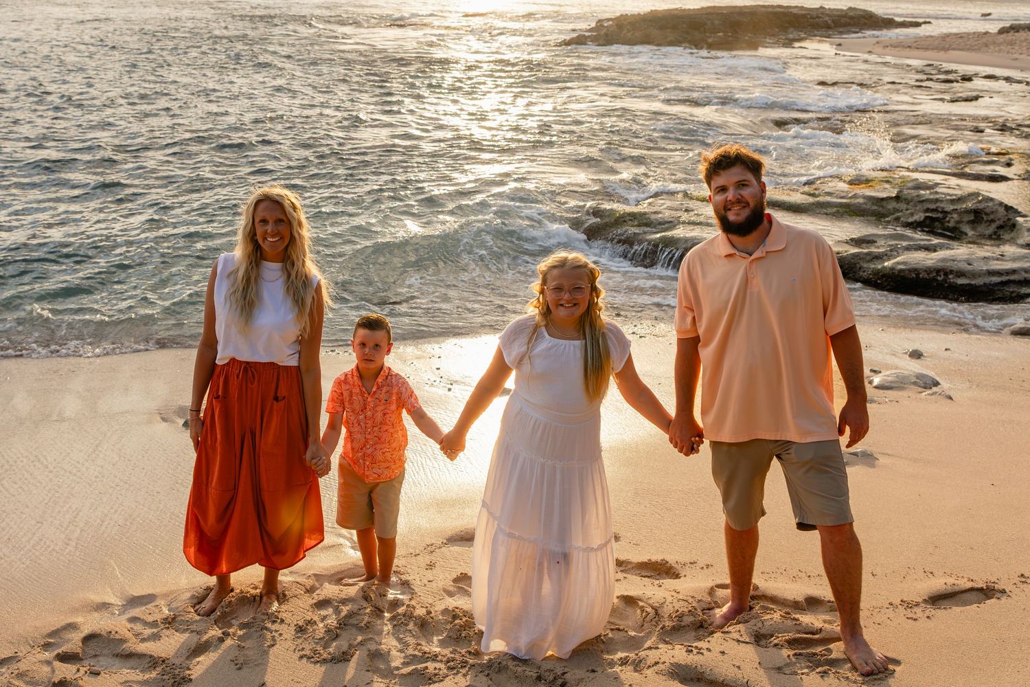 Family of four holding hands on a sandy beach at golden sunset with rocky shoreline in background.
