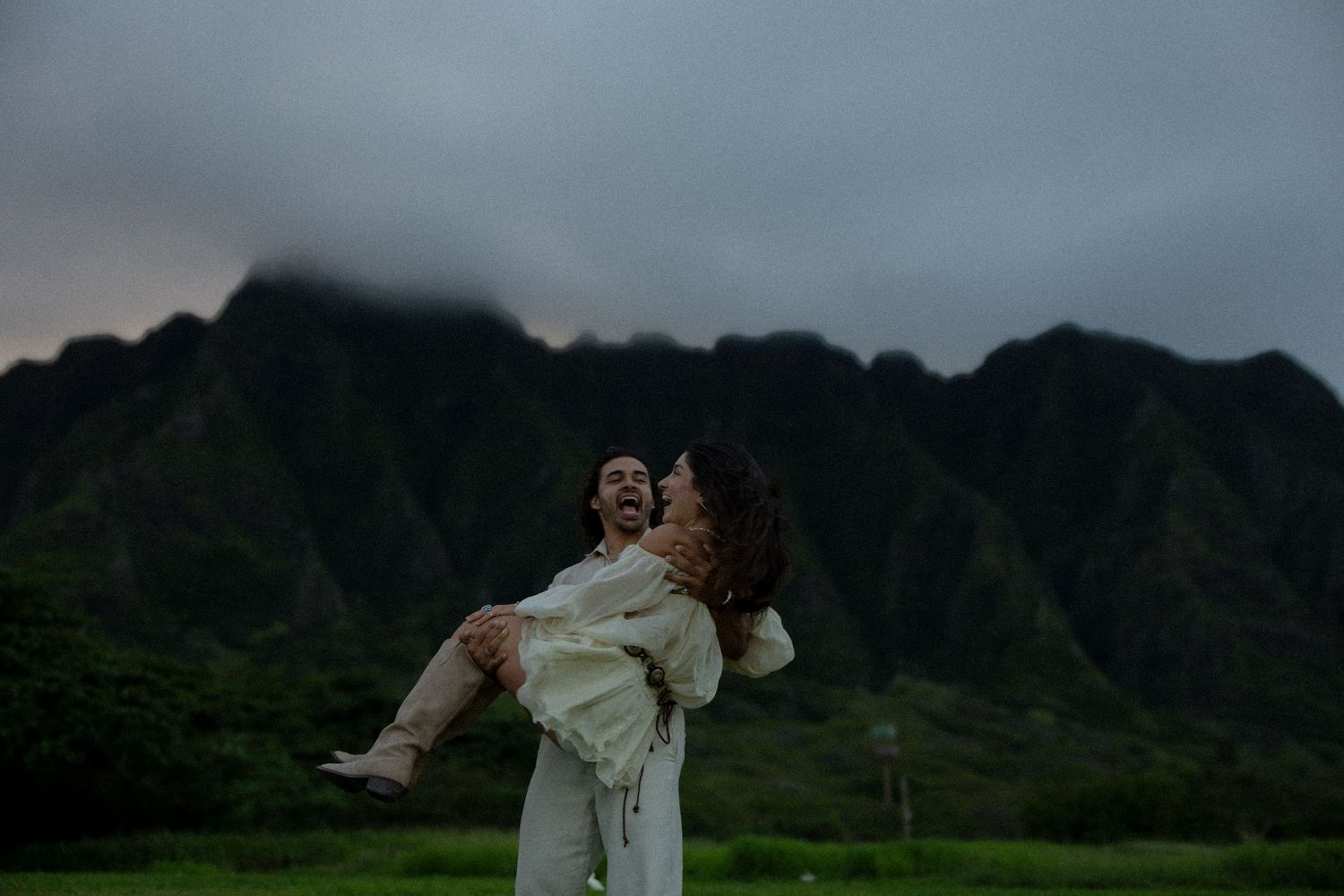 Couple embracing in front of lush green mountains under dramatic cloudy skies in Hawaii.