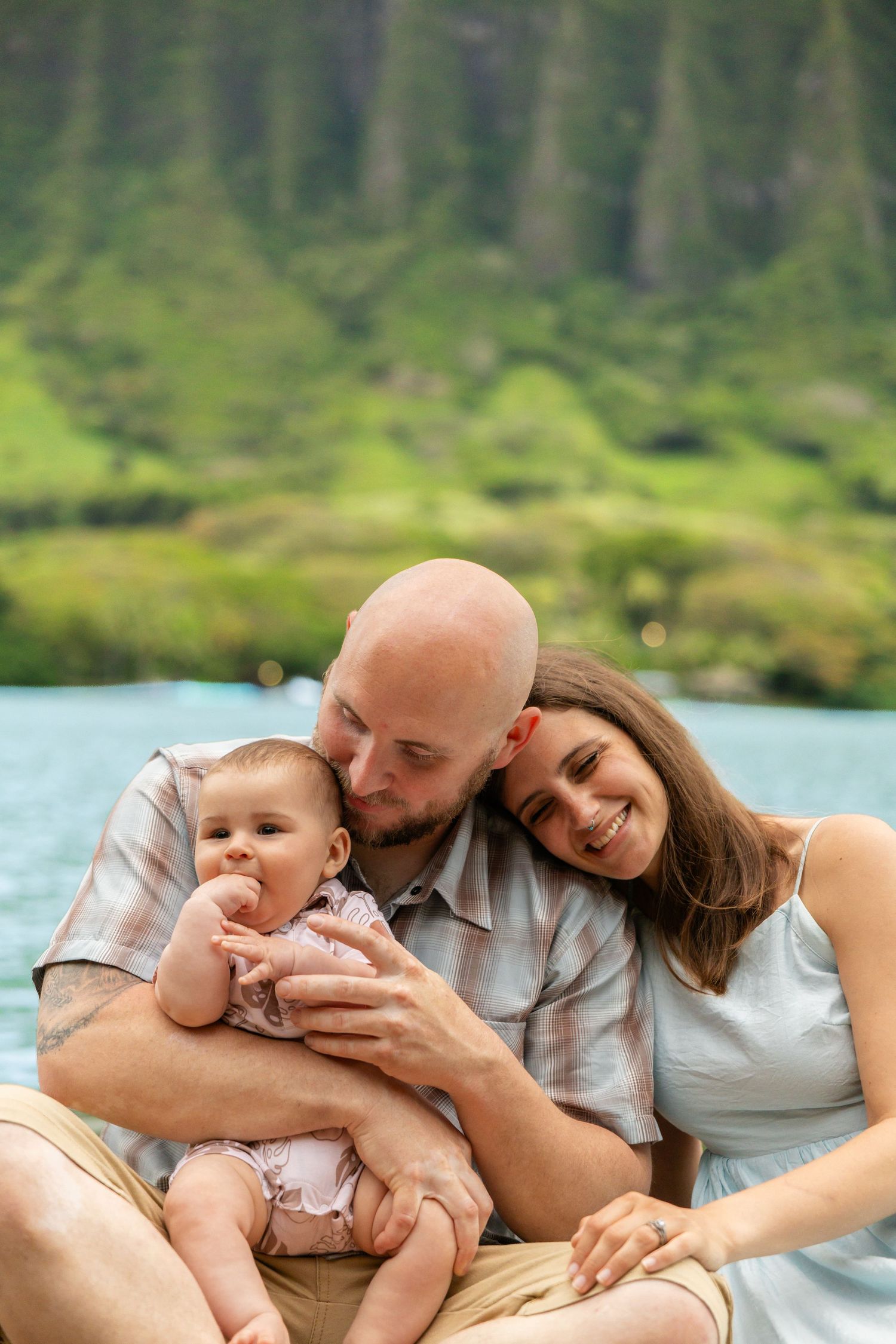 Happy family with bald father kissing baby while mother smiles near lush green mountains.