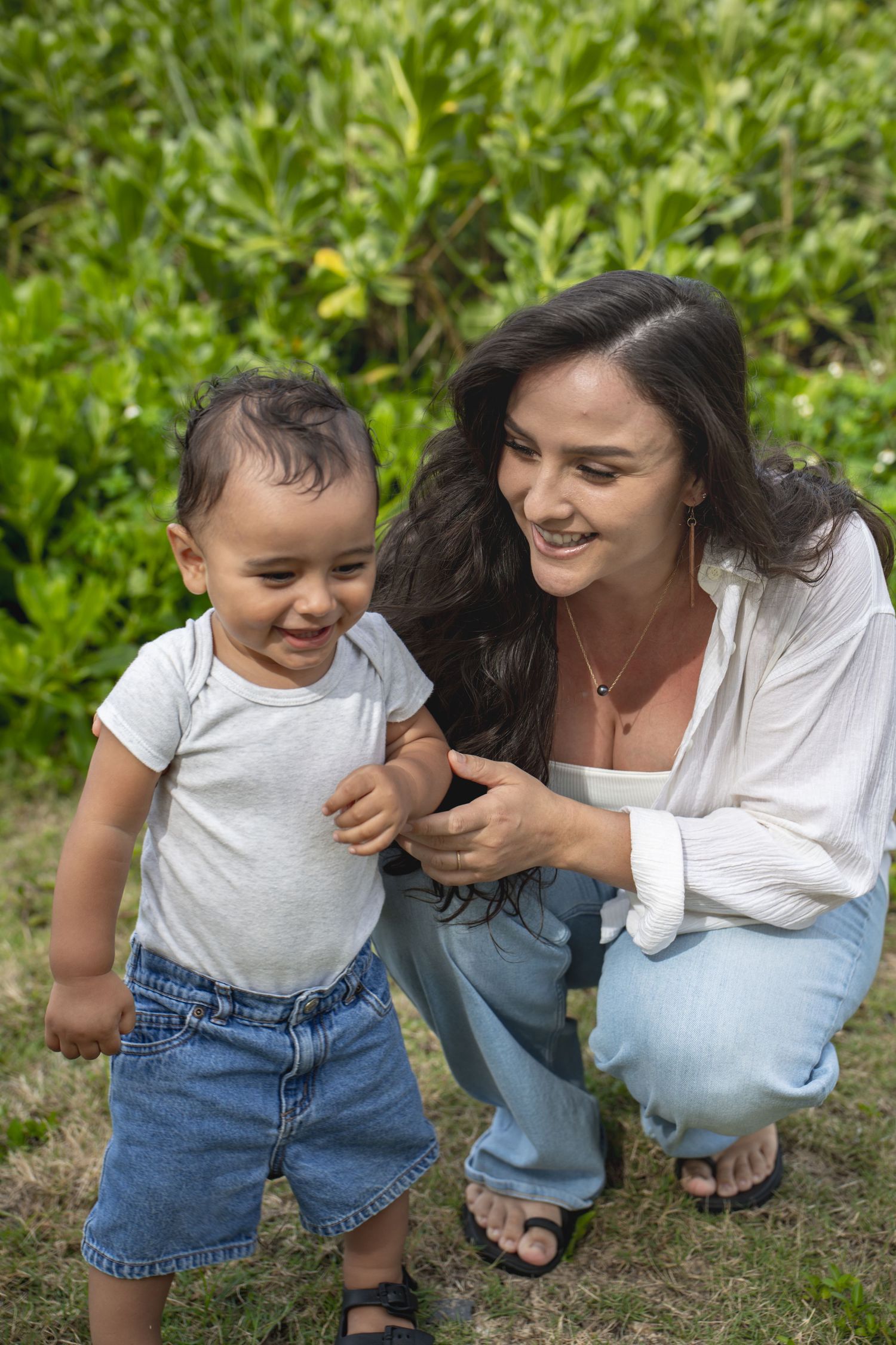Smiling young mother crouches beside toddler boy in garden setting.