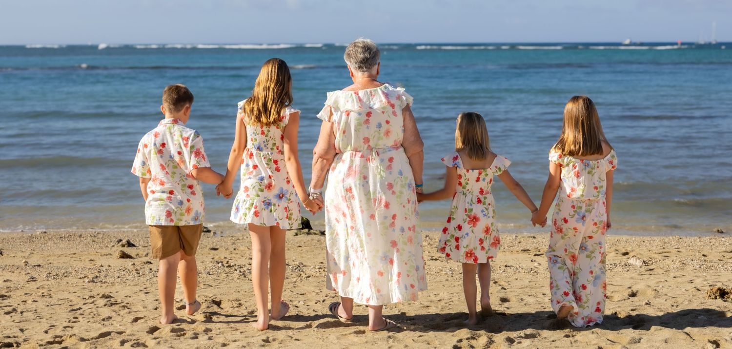 A mother and four children in matching floral outfits hold hands on a sandy beach with ocean waves in the background.